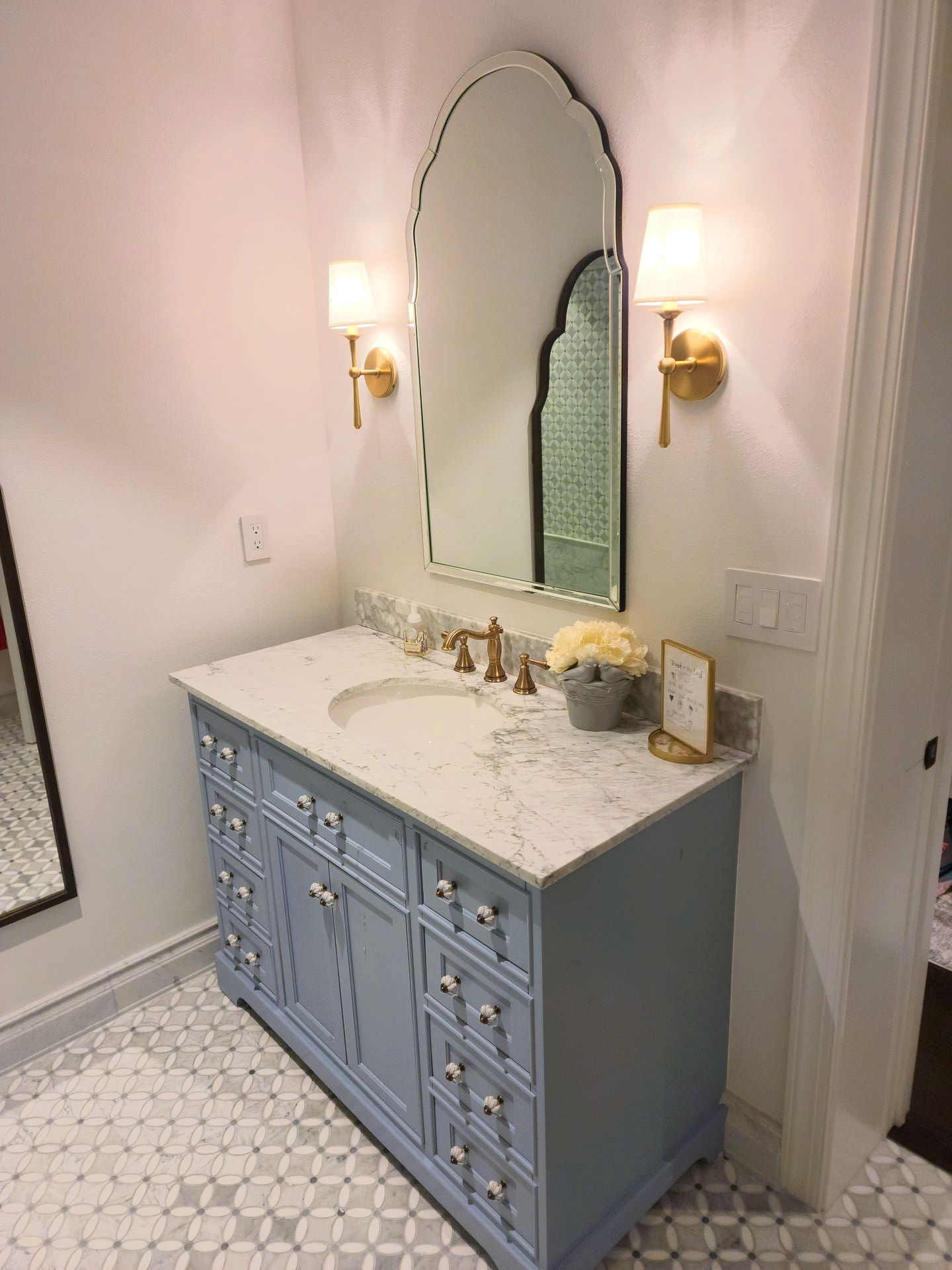 Light blue vanity with a white countertop, arched mirror, and gold sconces in a bathroom.