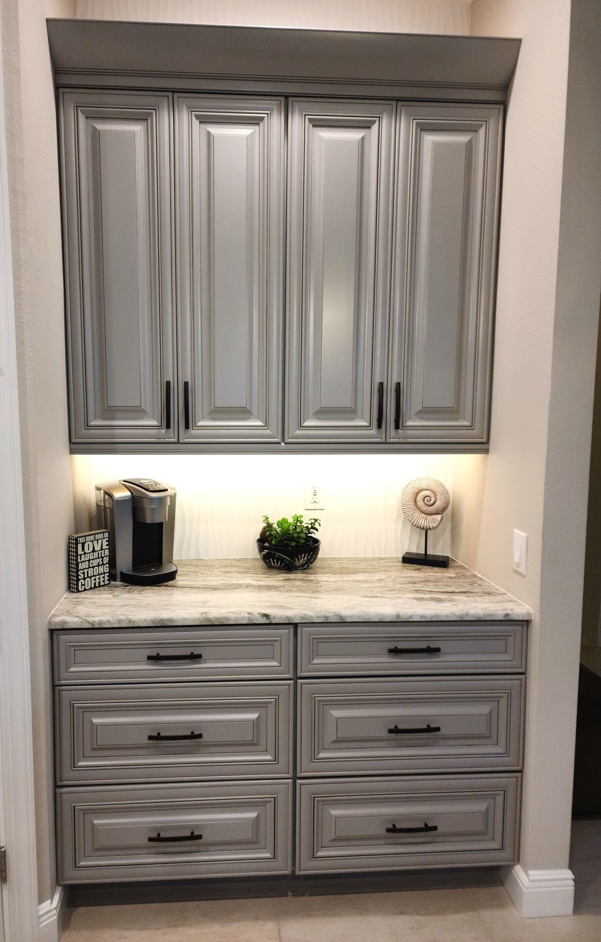 Grey cabinets with black hardware above and below a marble countertop, a coffee maker, and decor.