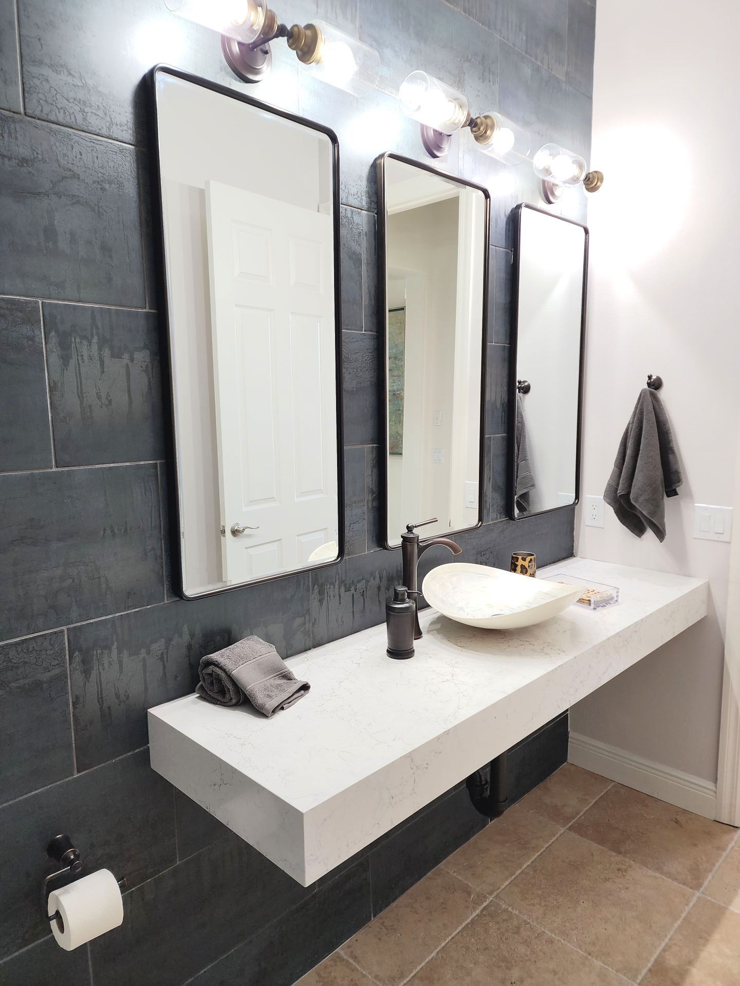 Modern bathroom with three black-framed mirrors, white floating vanity, dark gray tile wall, and a beige tiled floor.