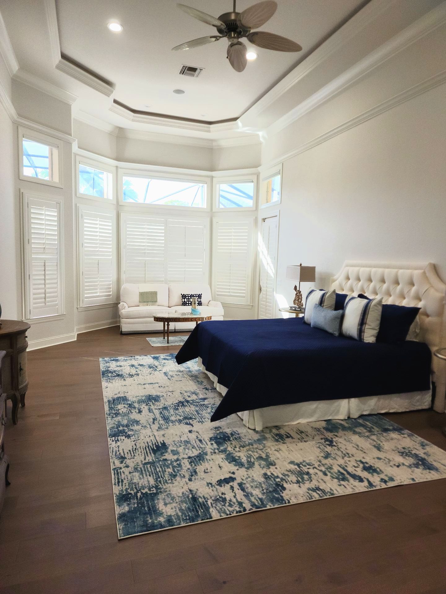 Spacious bedroom with a blue and white rug, white sofa, bay windows, and a bed with blue bedding.
