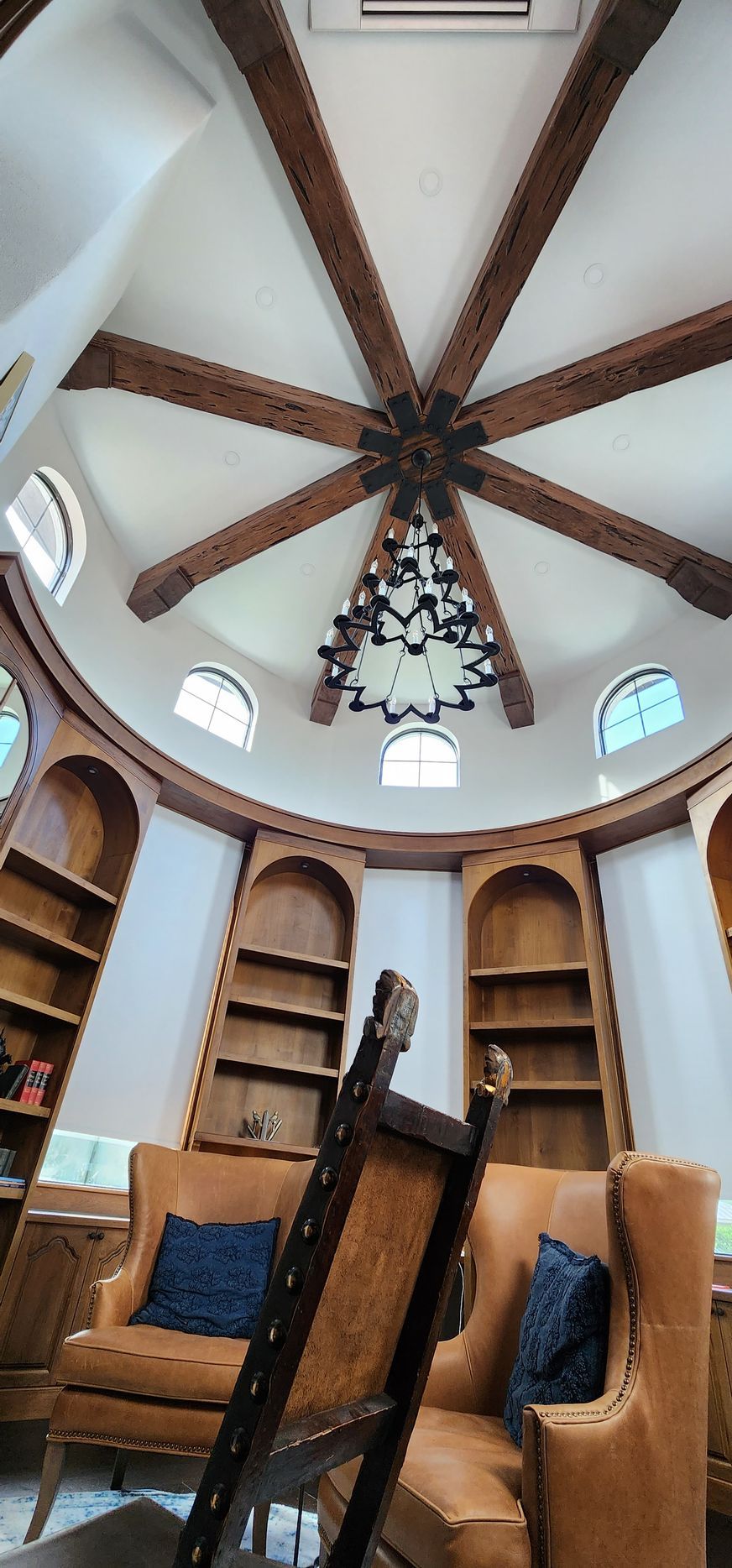 A circular library with wooden beams, arched bookshelves, armchairs, and a chandelier.