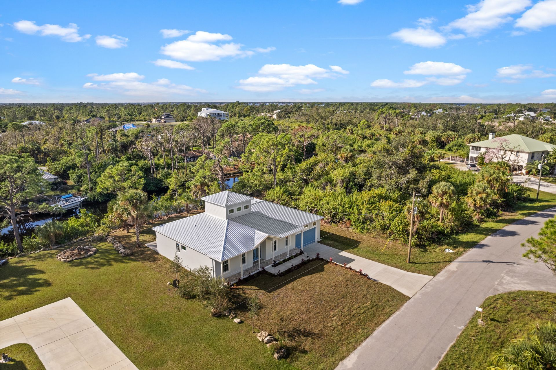 Aerial view of a white house with a metal roof, surrounded by lush green trees and a blue sky.