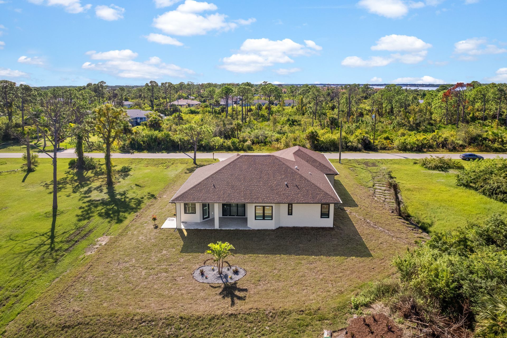 Aerial view of a white house with a brown roof, surrounded by trees and green grass under a blue sky.