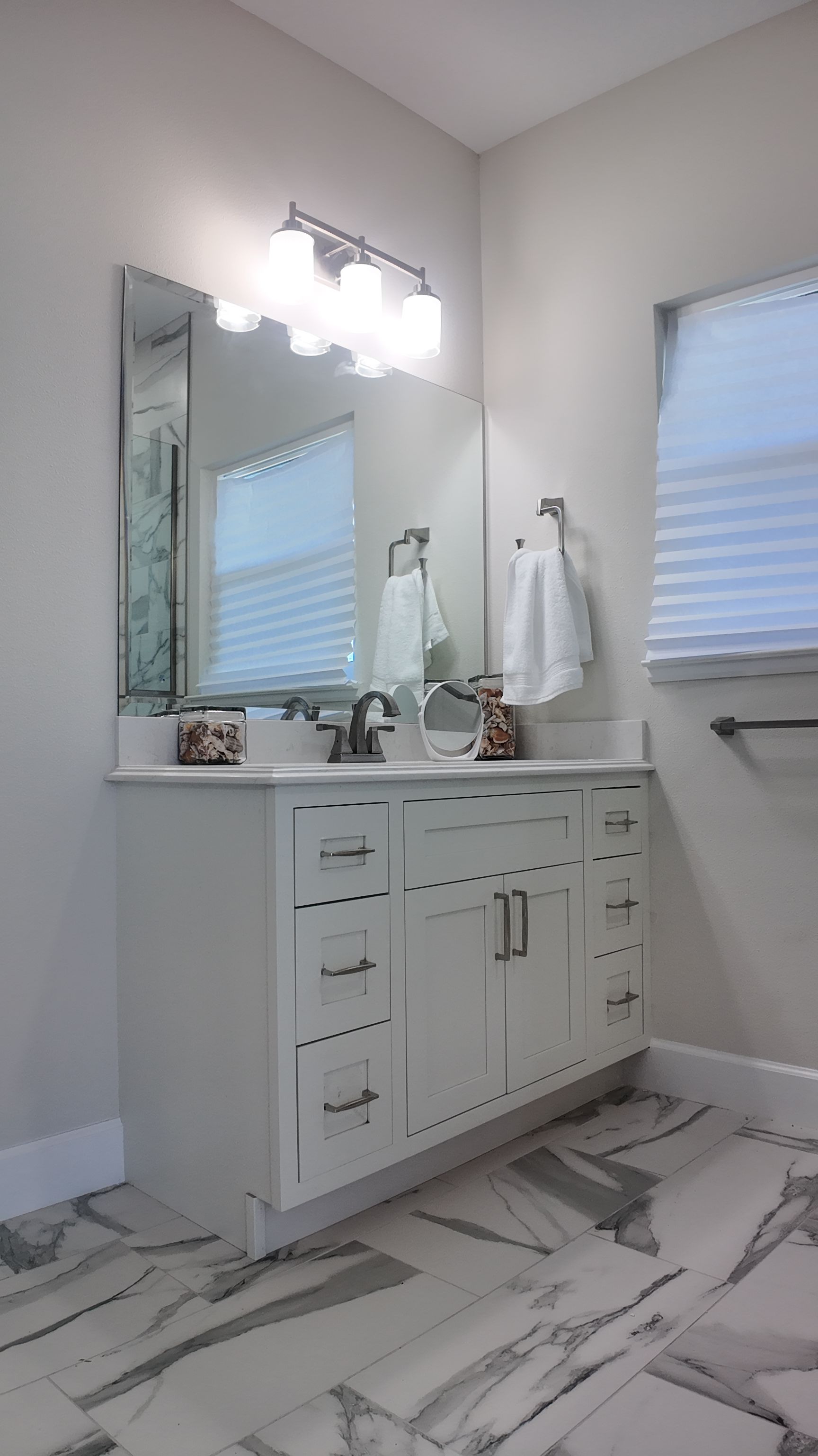 White bathroom vanity with mirror and light fixture, next to a window with blinds.