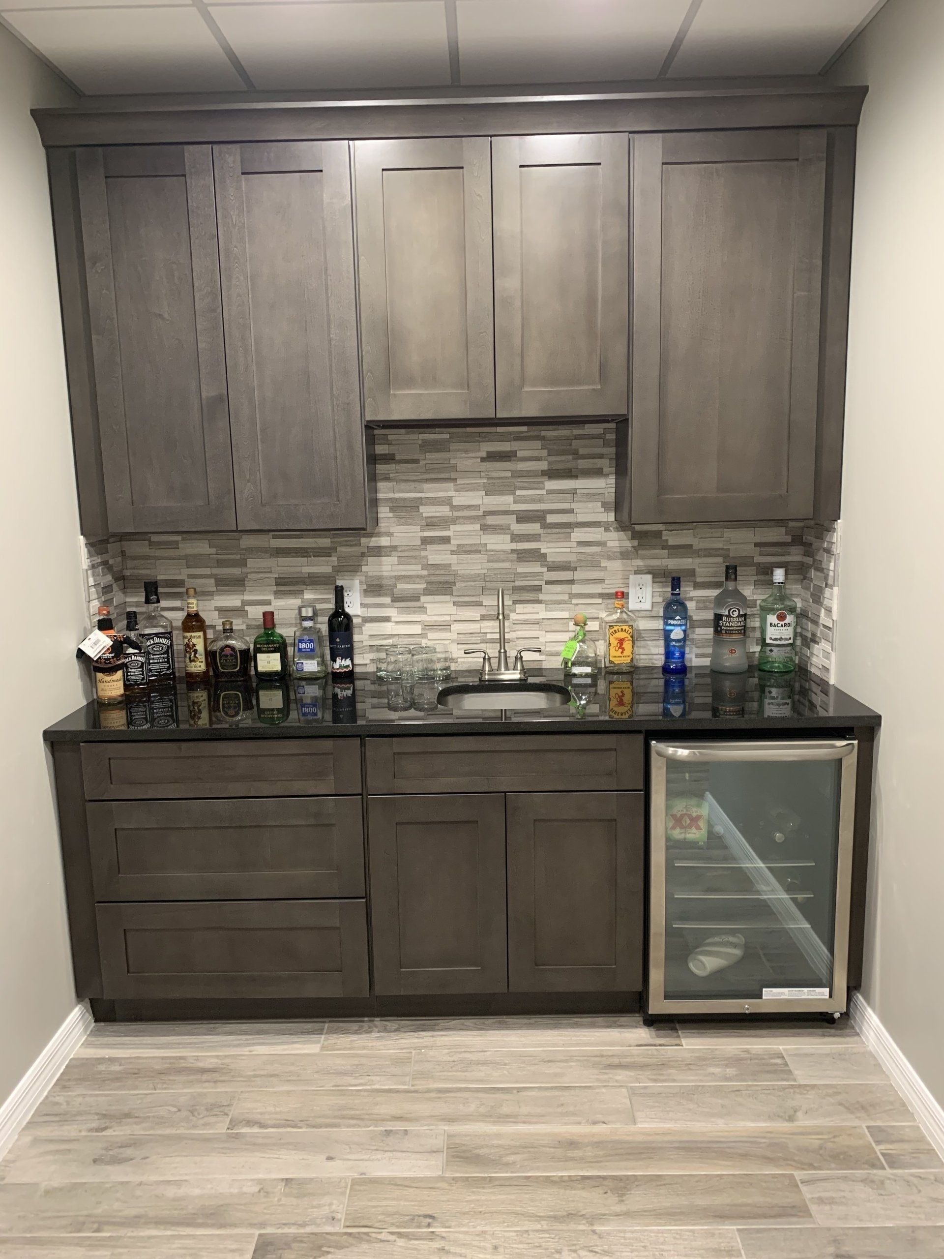 Gray bar cabinet with liquor bottles, mini-fridge, and sink against a backsplash and light-colored wall.