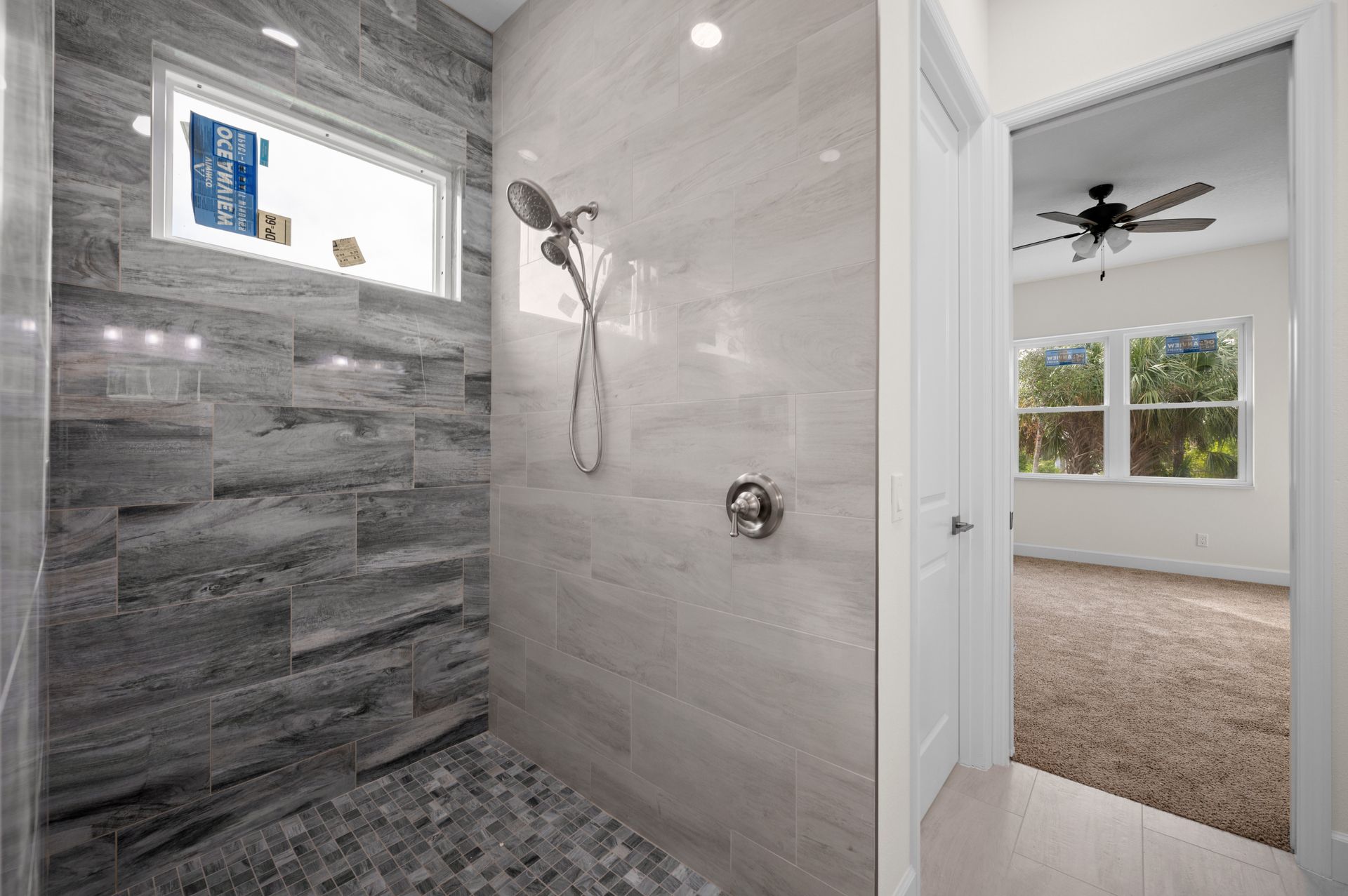 Shower with gray tile and pebble floor, adjacent to a bedroom with a ceiling fan.