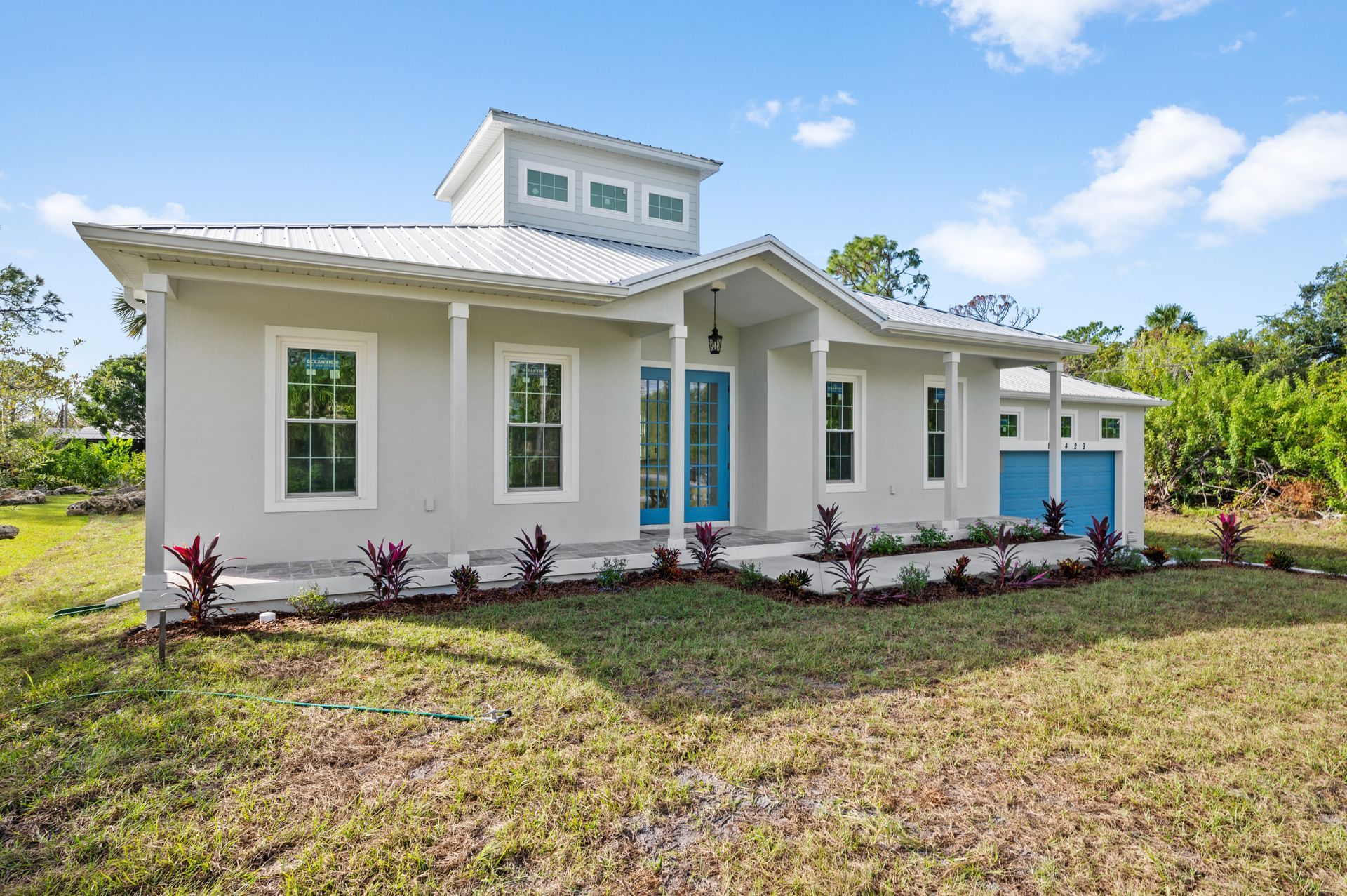 Gray stucco house with teal doors and trim, white columns, and a rooftop tower; set in a grassy yard.