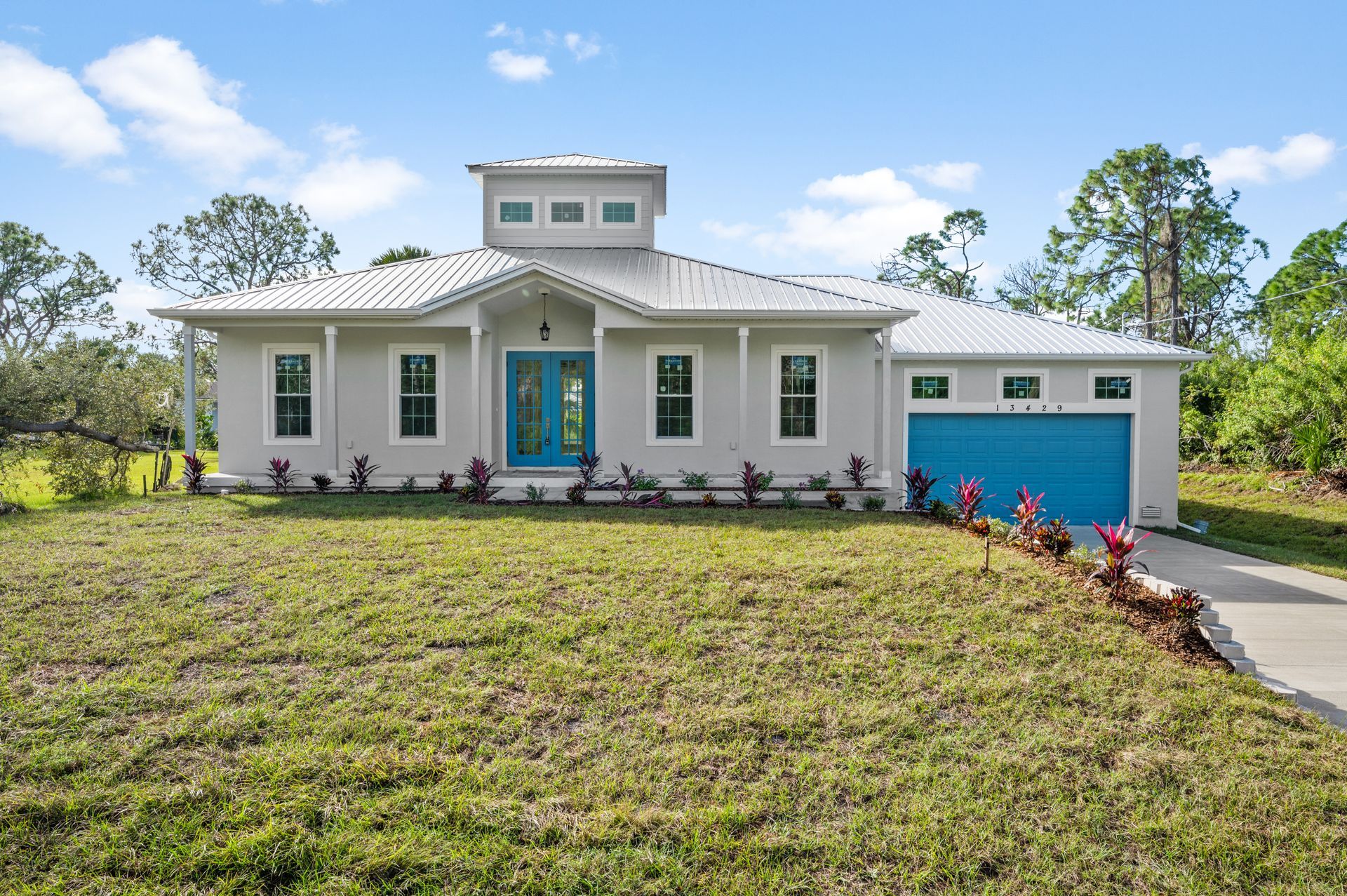 Light gray house with teal doors/garage; grassy yard, surrounded by trees under a blue sky.