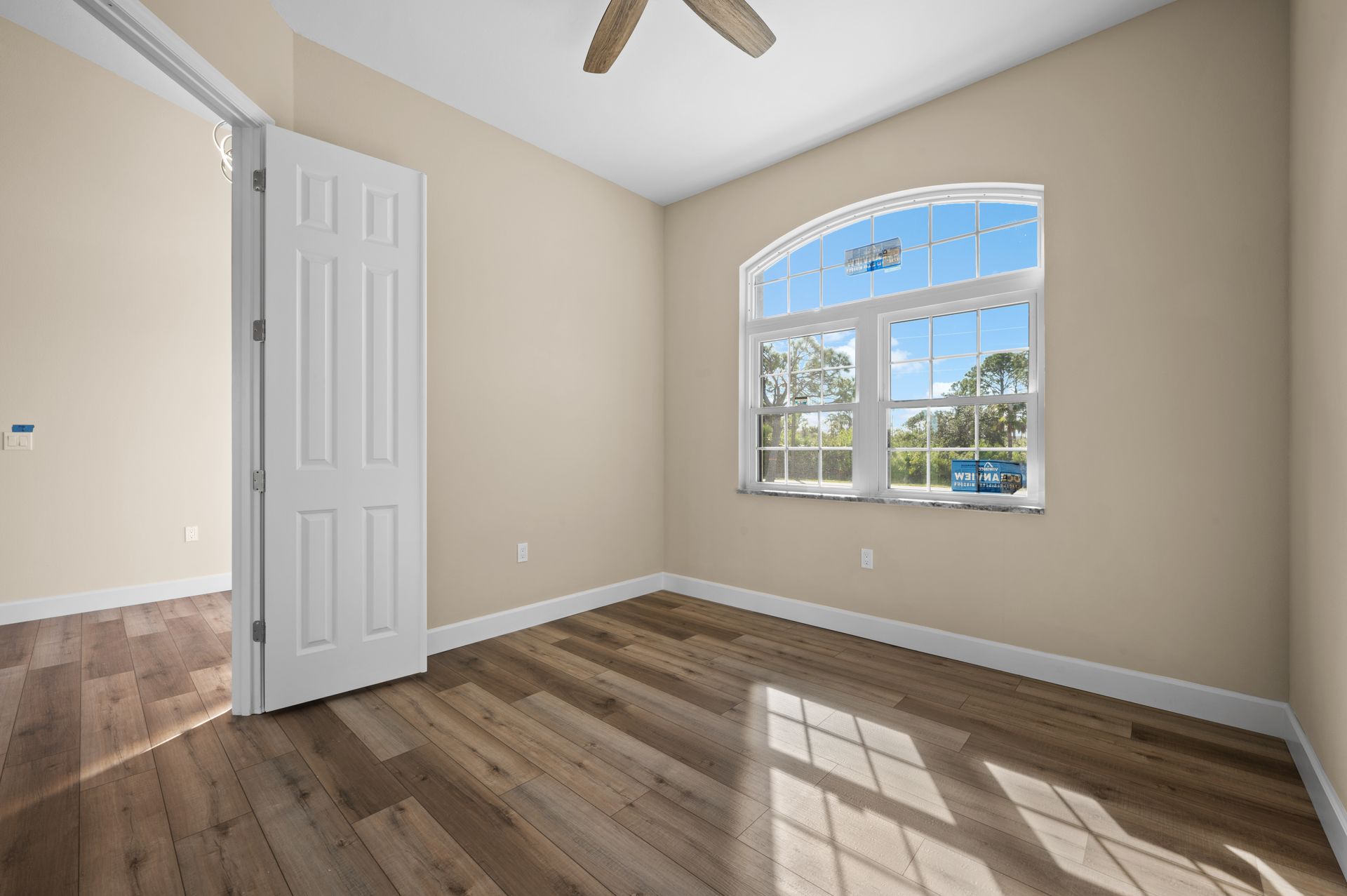 Empty bedroom with wooden floors, arched window with sunlight, and open doorway.