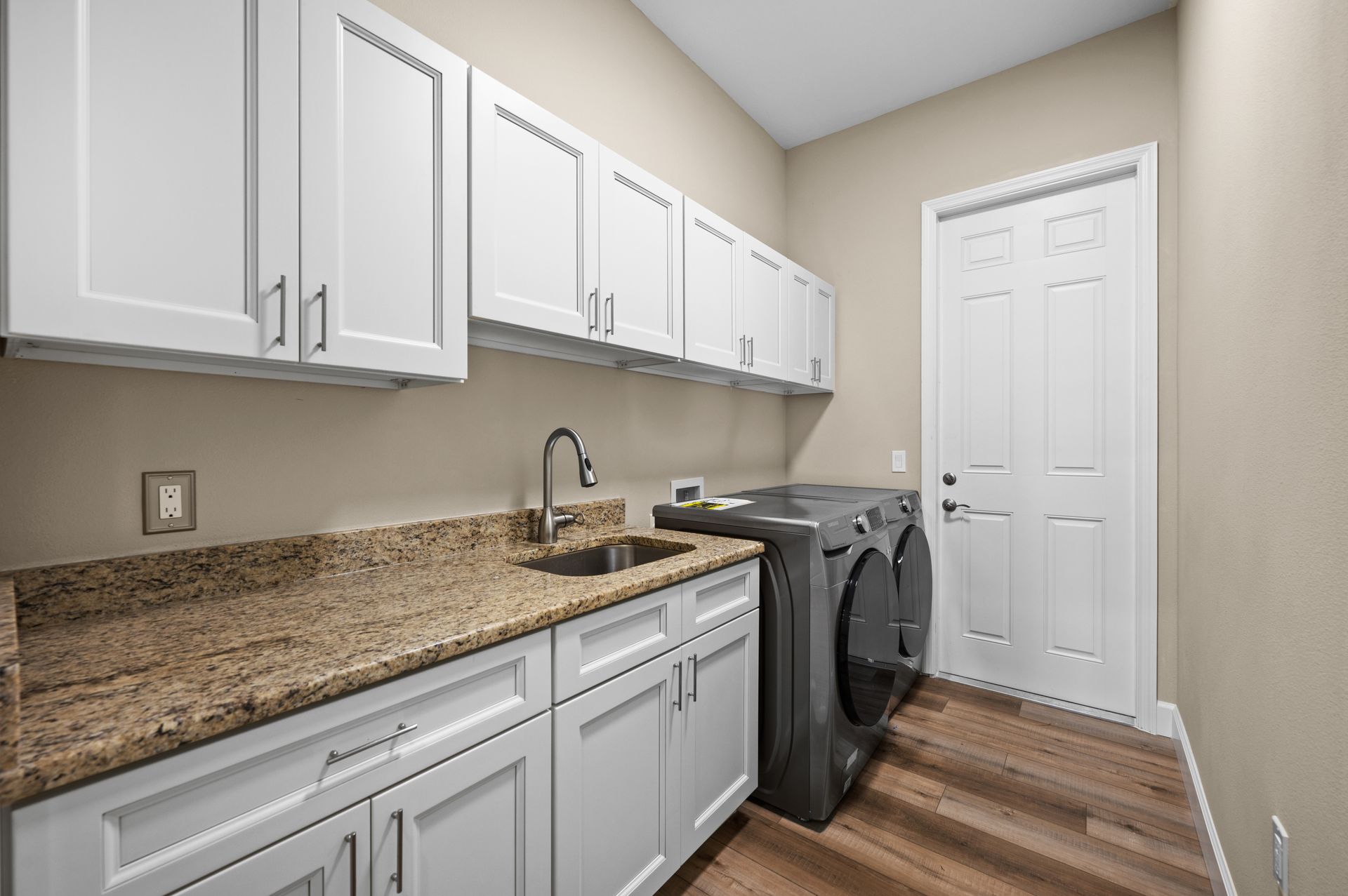 Laundry room with white cabinets, a sink, washer/dryer, and a door. Tan walls and wood-look flooring.