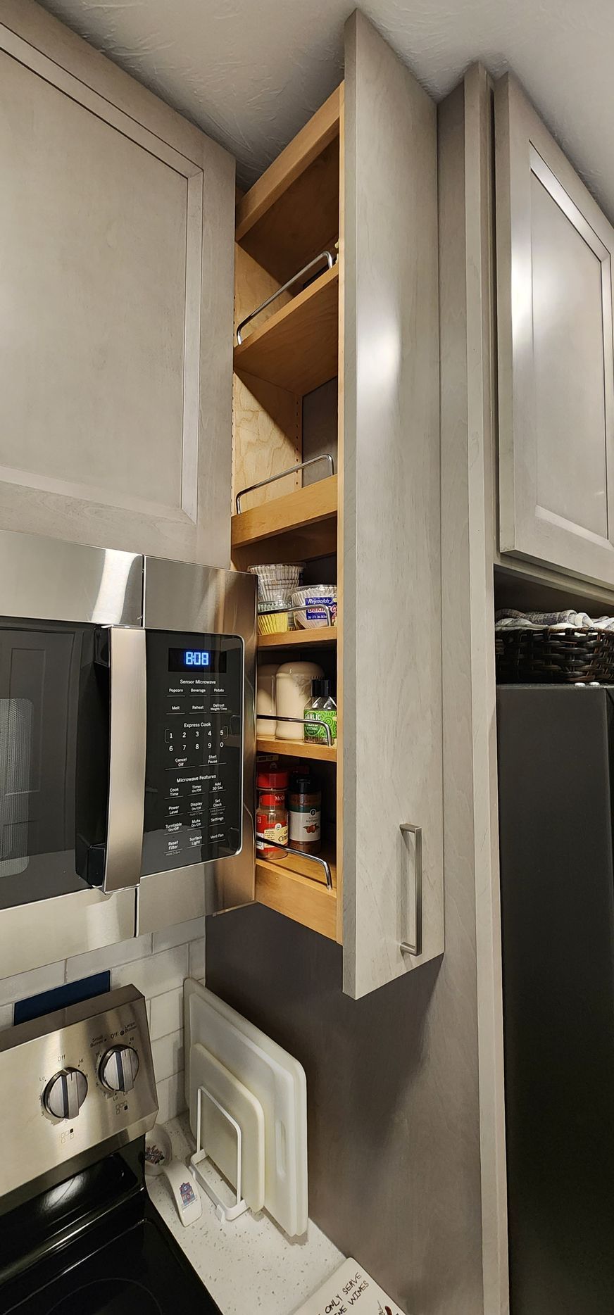 A tall, narrow pull-out cabinet with shelves in a kitchen, filled with spices and food items, next to a microwave.