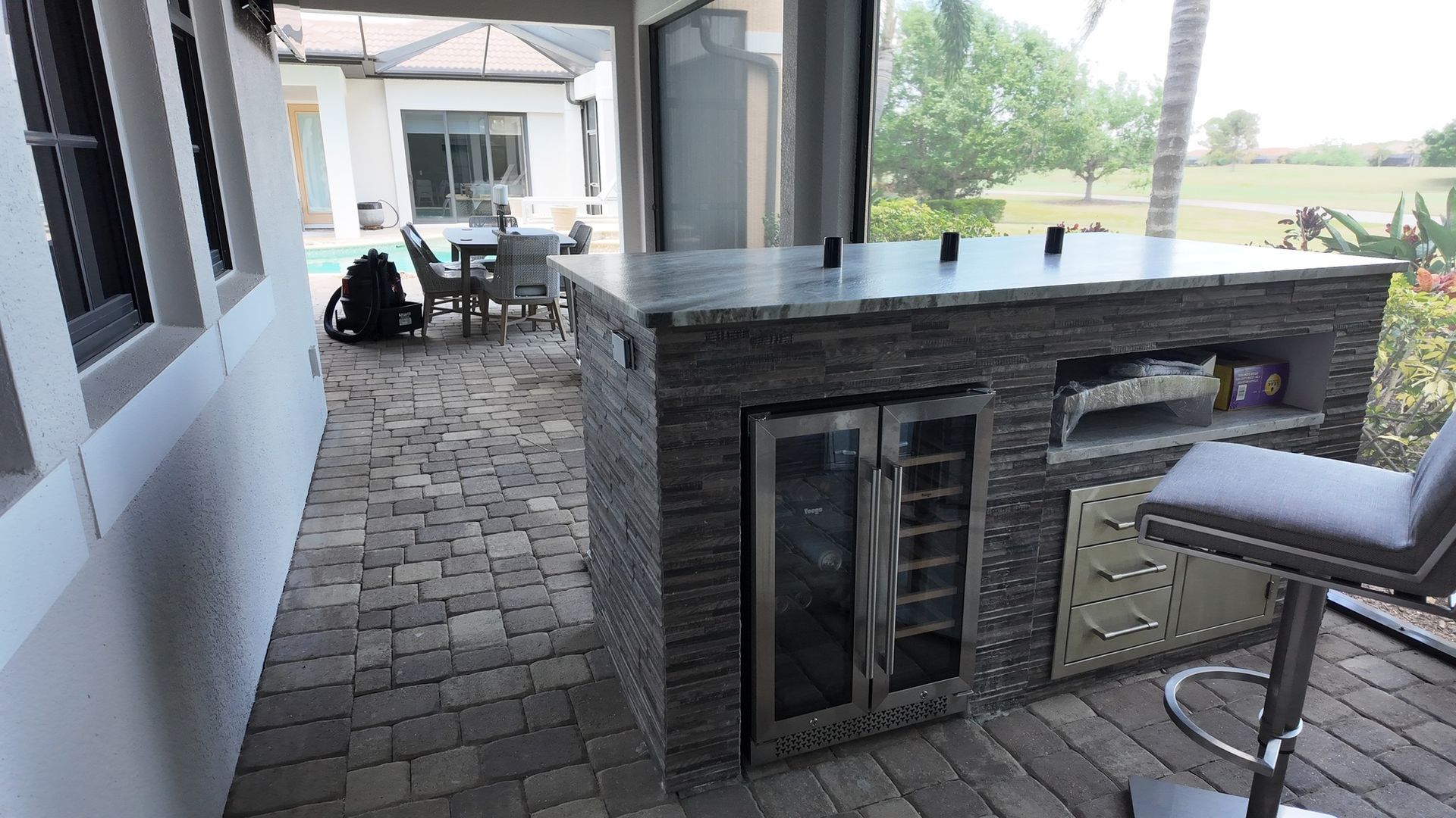 Outdoor kitchen with stone exterior, bar seating, a built-in fridge, and grill, overlooking a patio and yard.