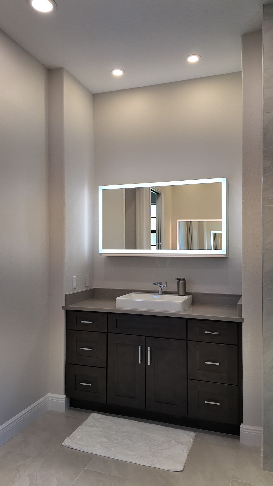 Bathroom with dark brown vanity, white sink, illuminated mirror, and light gray walls.