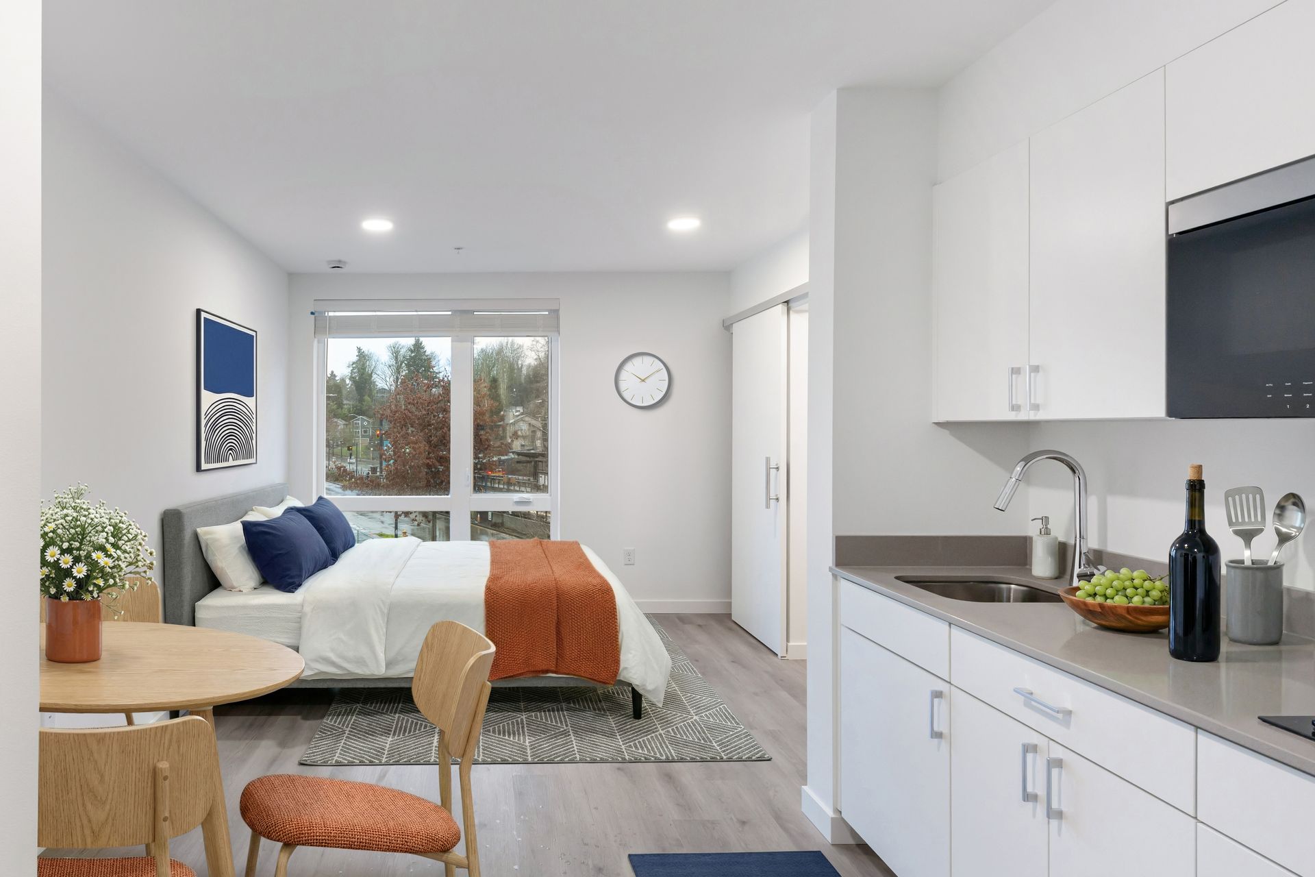 Studio apartment with bed, small kitchen, round table, and a window overlooking trees.