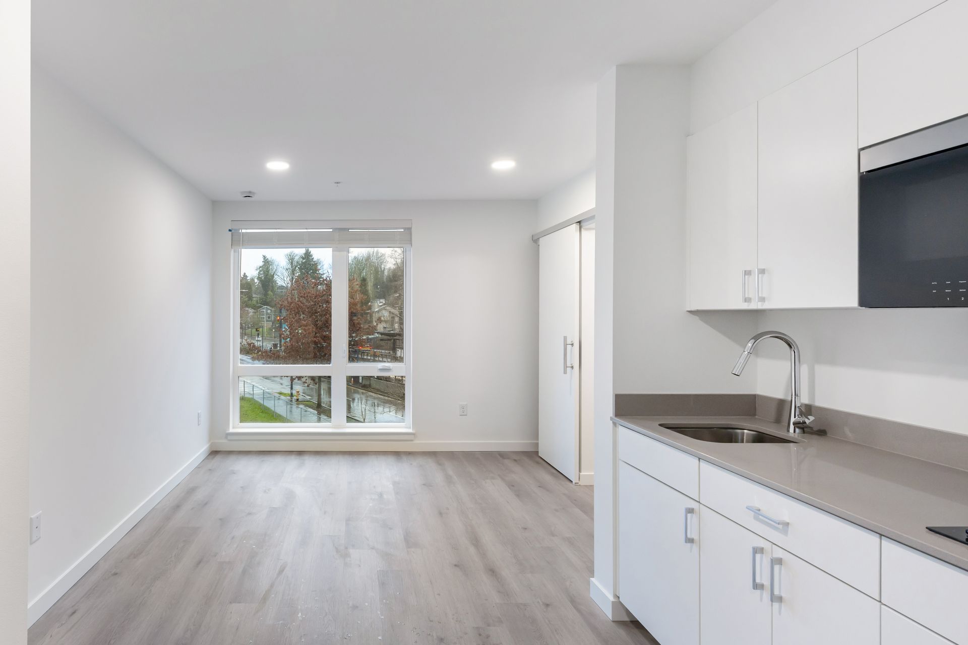 A bright, empty studio apartment with a small kitchen area and large window.
