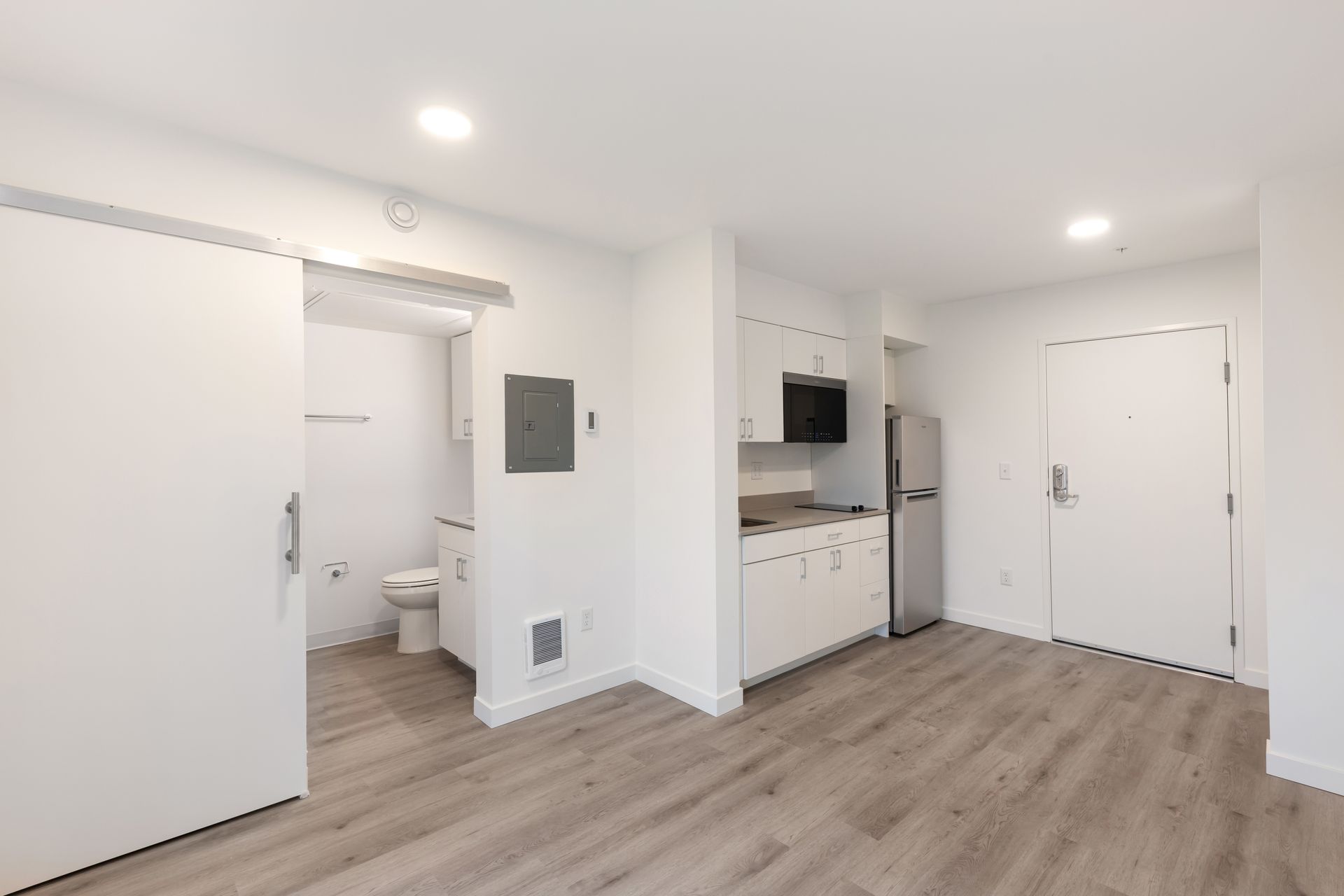 Interior of a studio apartment with a bathroom, kitchen, and light wood flooring.