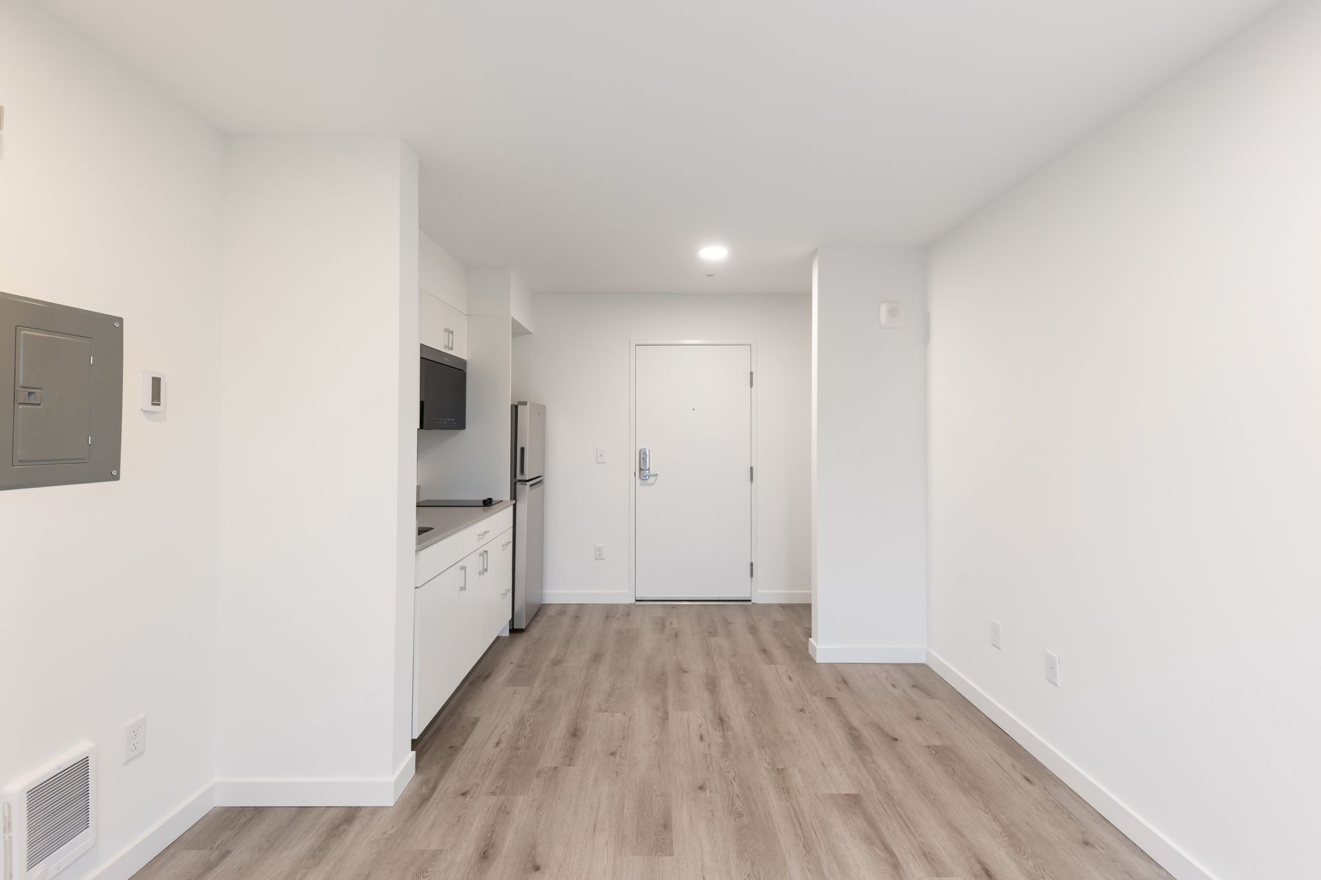 Empty white studio apartment with light wood flooring, kitchen in the back, and a closed door.