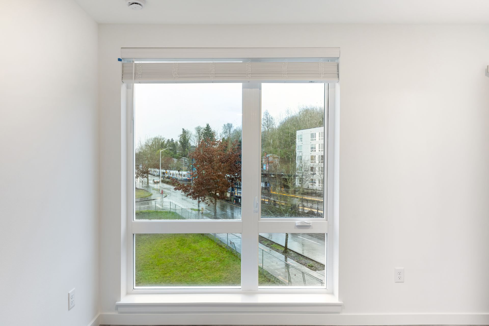 Window with a view of a rainy street, a building, trees, and a green lawn; white walls and a shade.