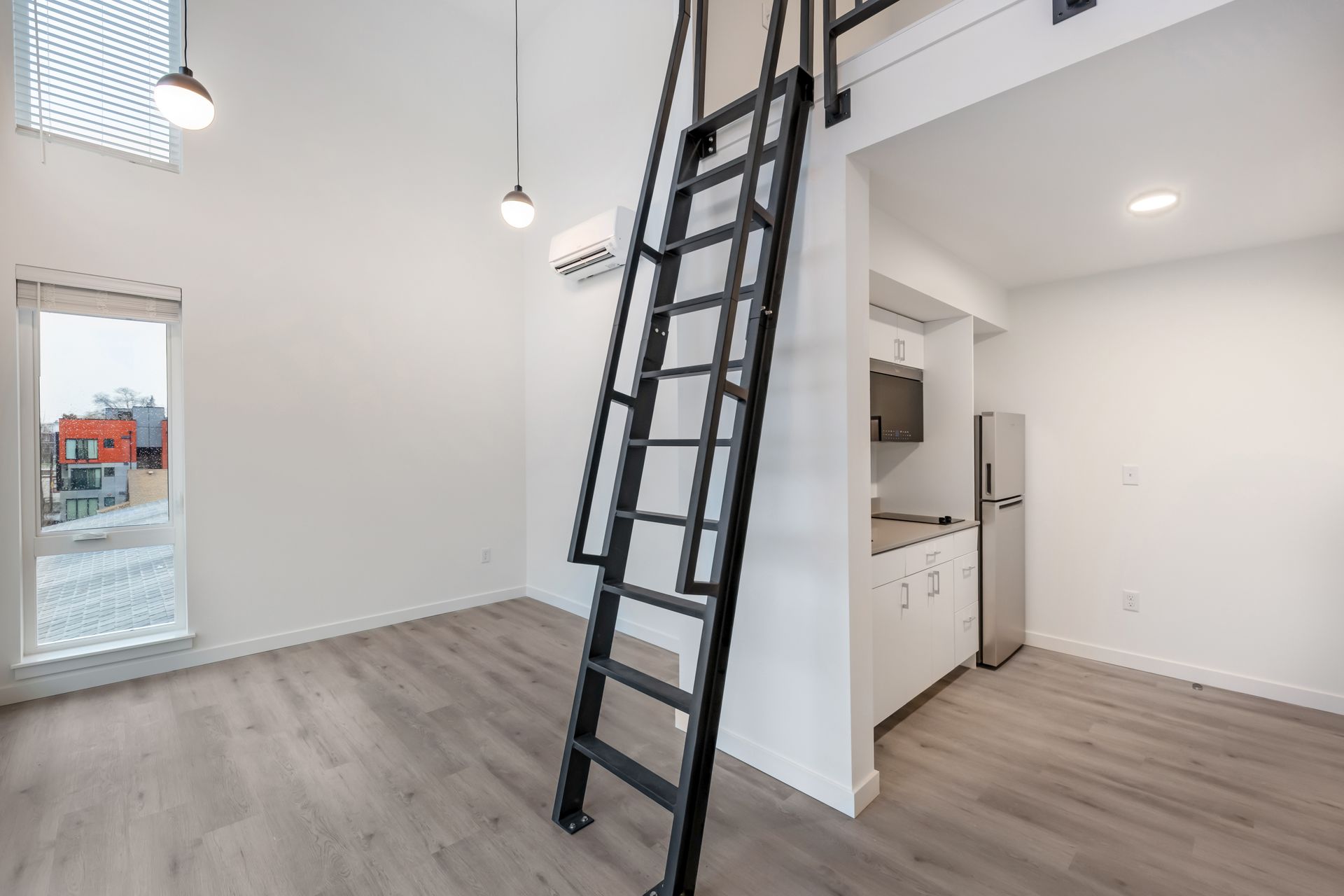 Interior of a modern, white apartment with a ladder to a loft, small kitchen, and large window.