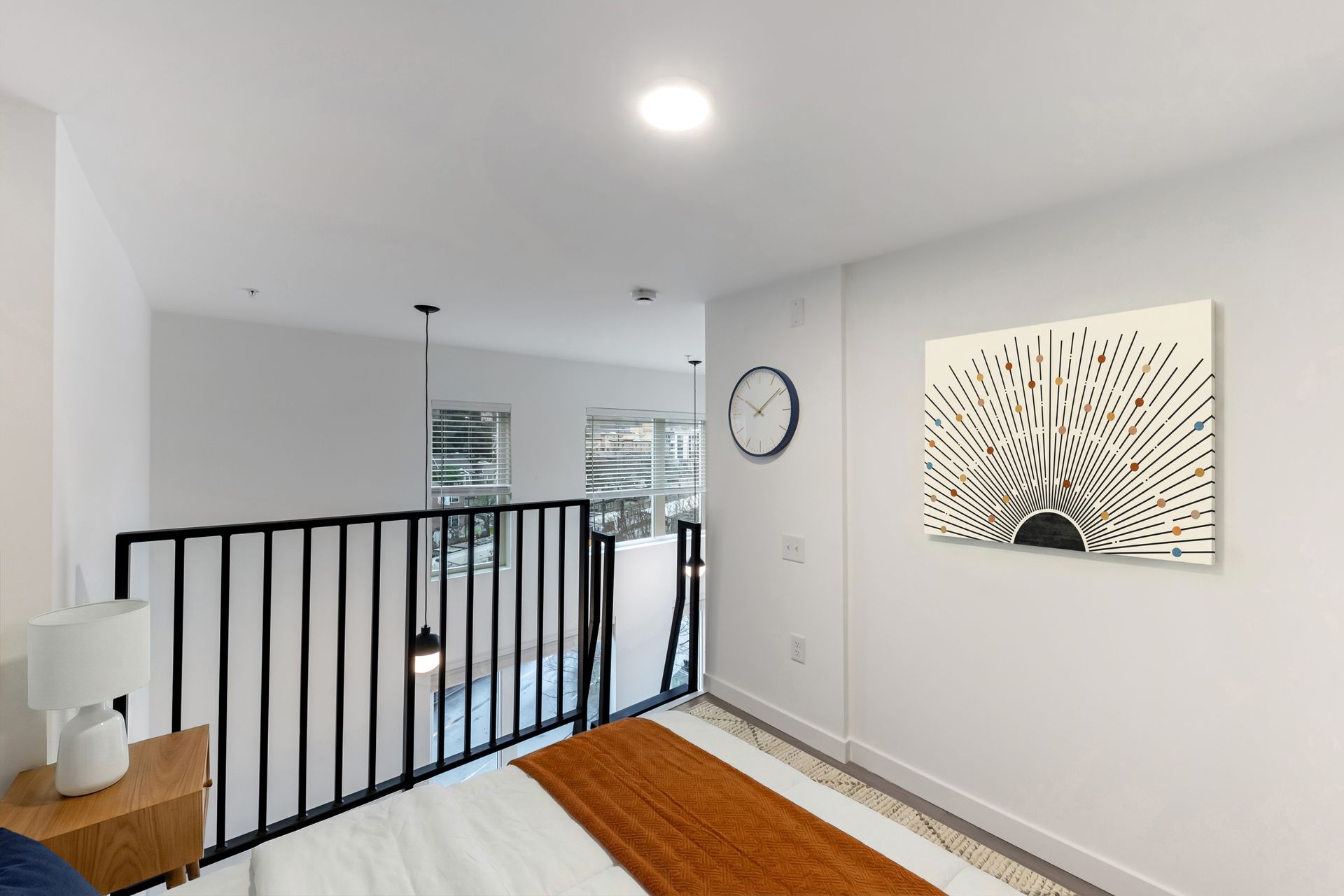 Loft bedroom: white walls, black railing, bed with orange blanket, clock, abstract art, pendant lights.