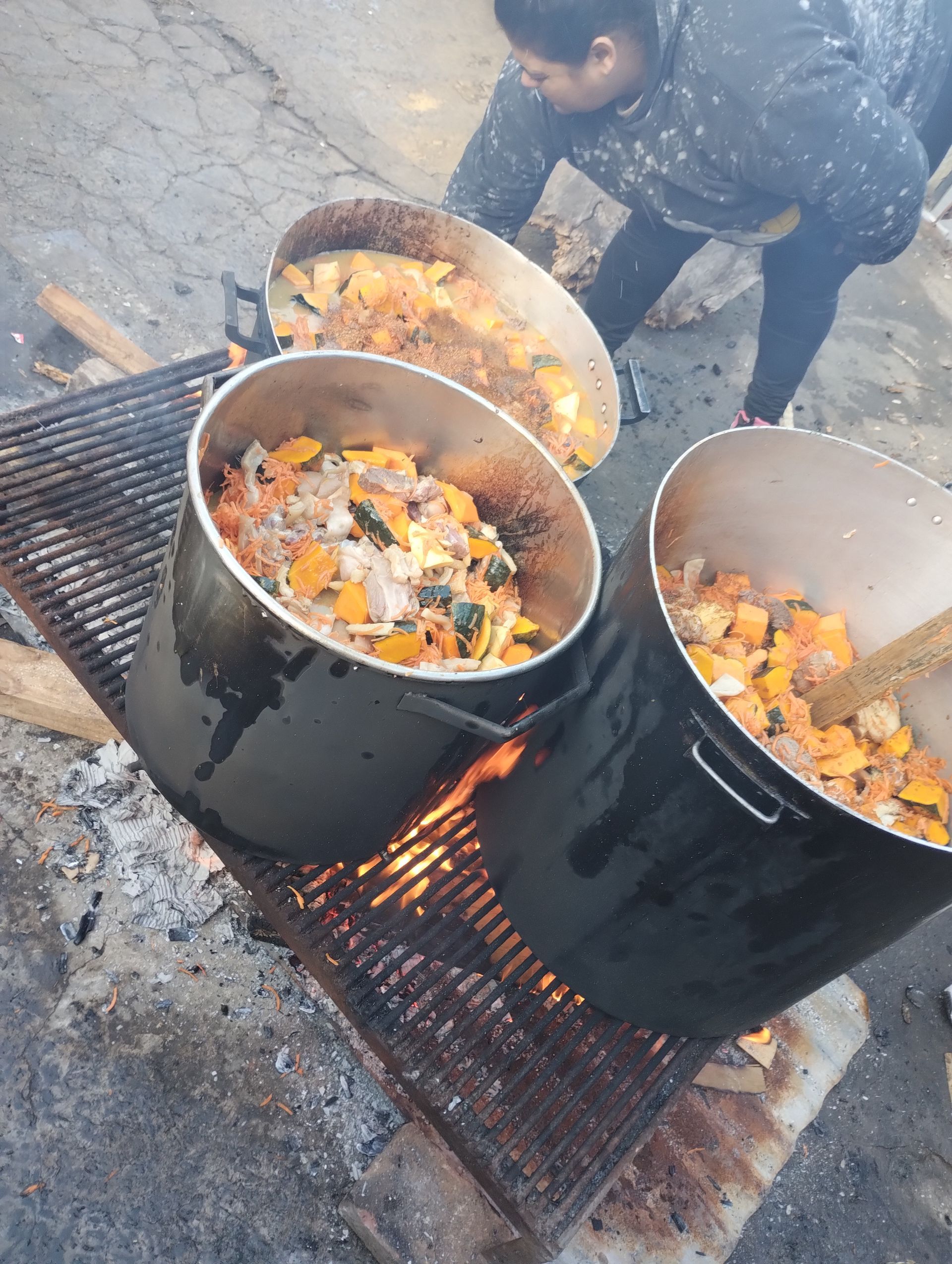 Ollas de comida cocinándose sobre el fuego en una parrilla; una persona vierte la comida desde un recipiente.