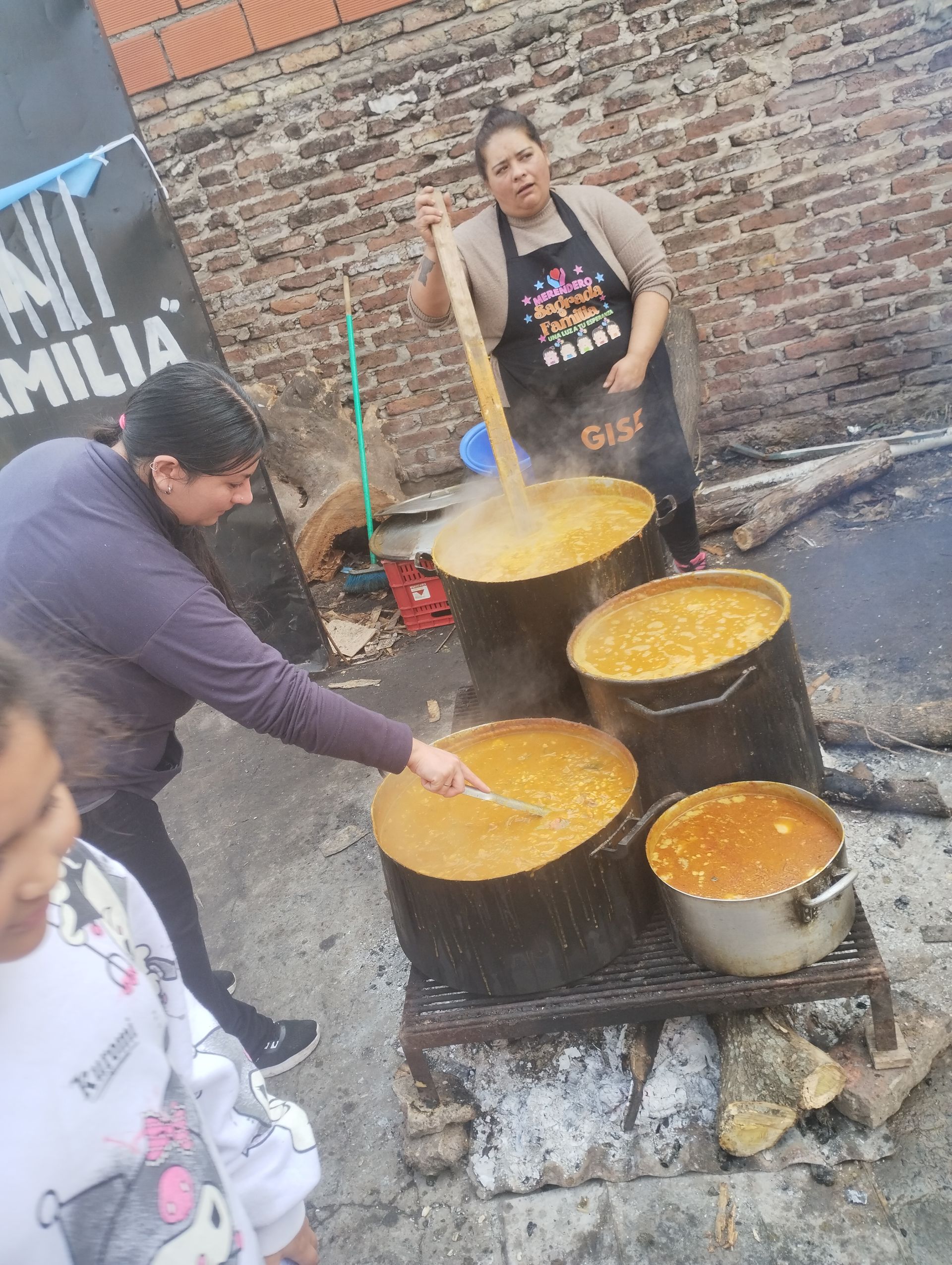 Personas revolviendo grandes ollas de comida sobre el fuego. Una mujer con delantal revuelve con una gran cuchara de madera.
