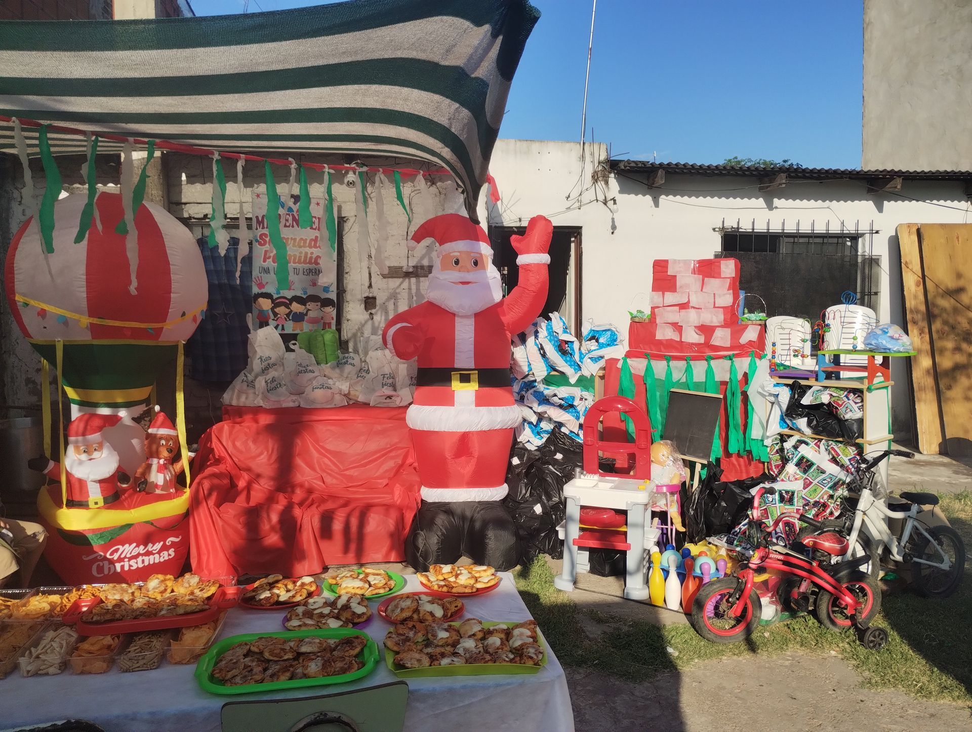 Una exhibición navideña en el exterior. Un Papá Noel inflable, comida y juguetes sobre una mesa.