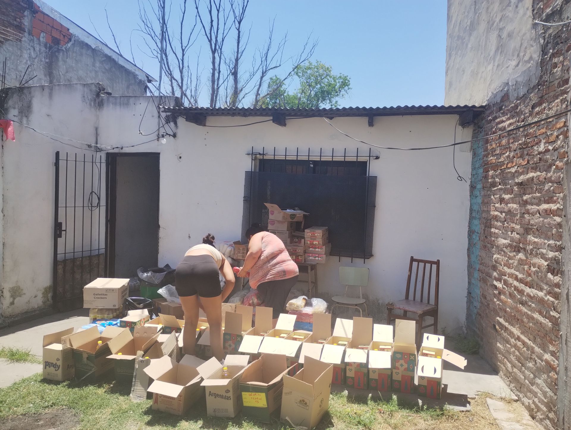 Una mujer y un niño guardan comida en una caja de cartón al aire libre. Hay otras personas al fondo.