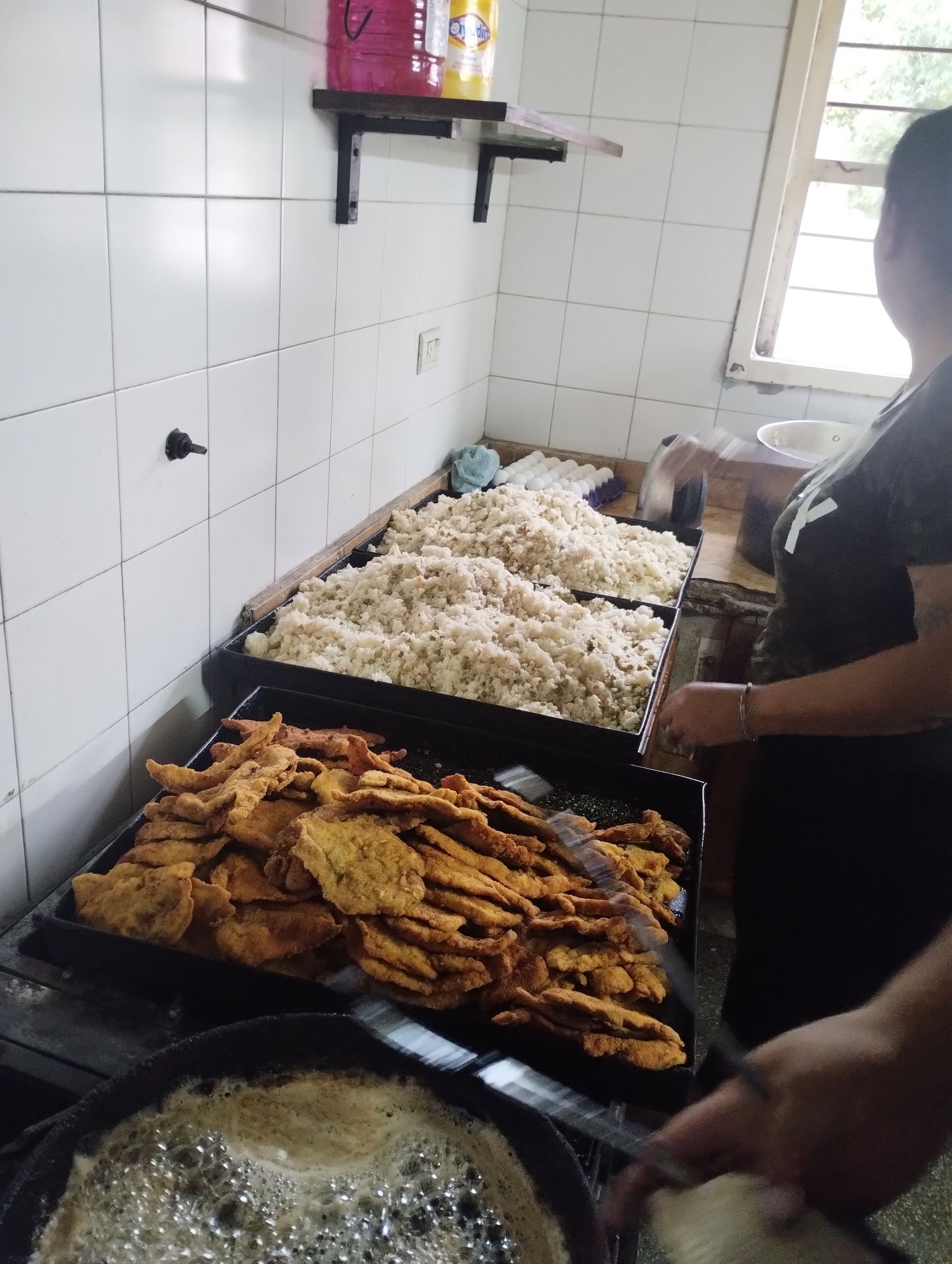 Una cocina con comida preparada en bandejas y una persona cocinando. Se ven frituras y arroz.