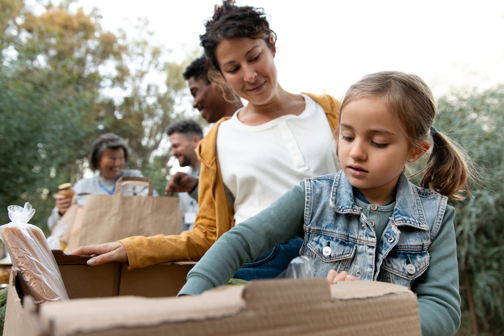 Una mujer y un niño guardan comida en una caja de cartón al aire libre. Hay otras personas al fondo.