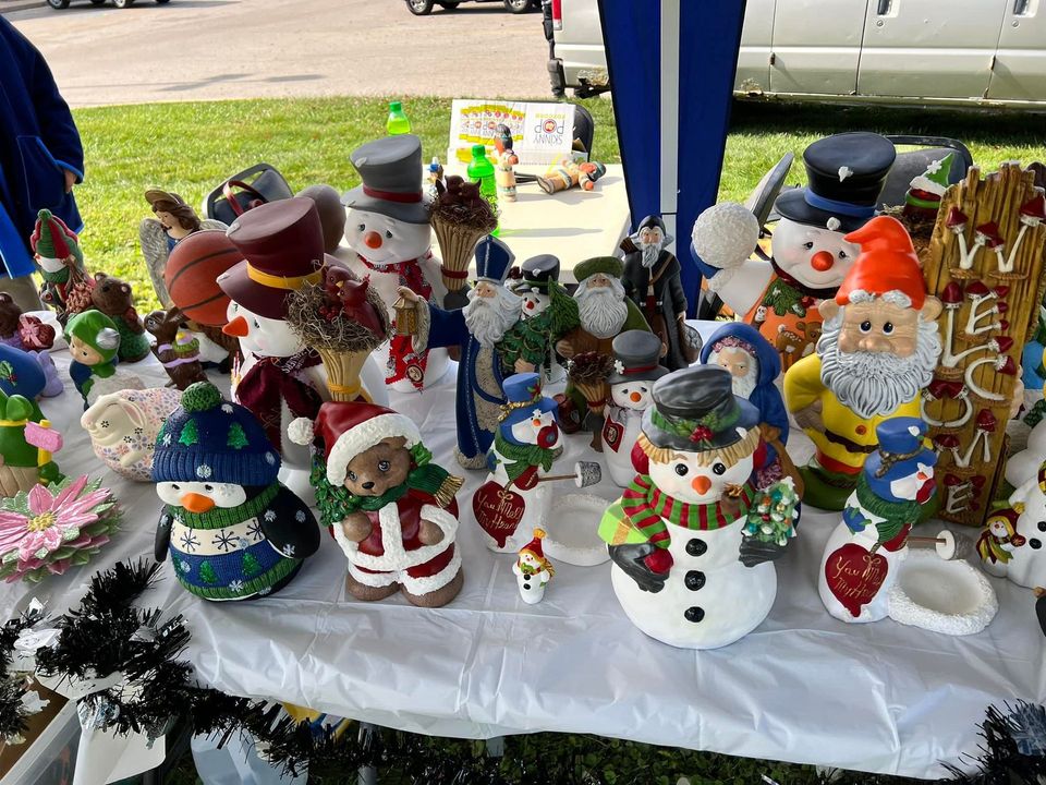 A table displaying various ceramic holiday figures, including snowmen and Santa, for sale outside on a sunny day.