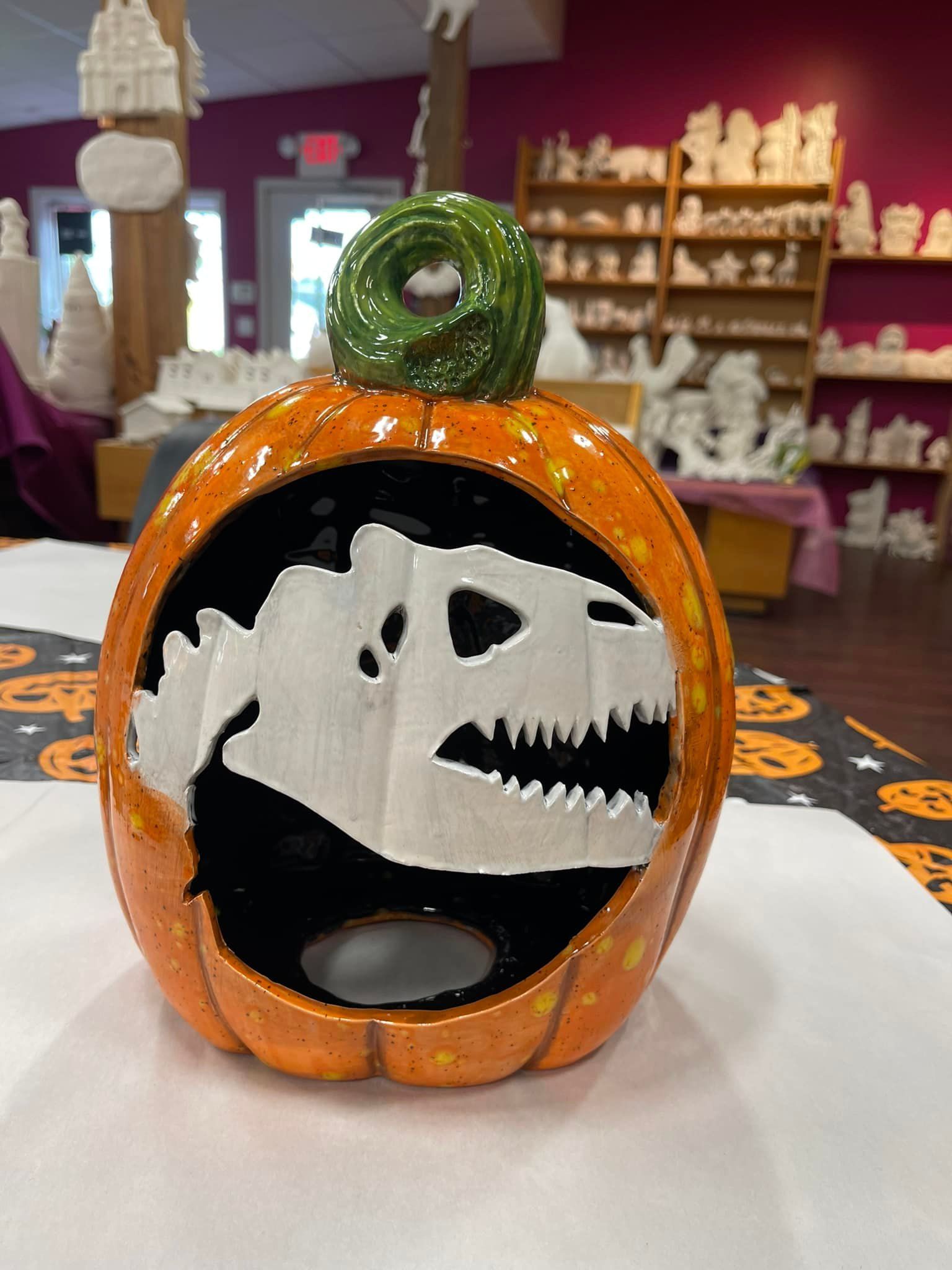 Ceramic pumpkin with T-Rex skull cutout, lit inside, on a table in a pottery studio.