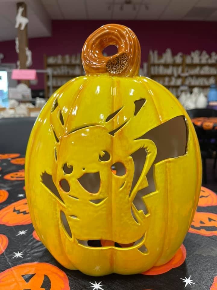 Yellow Pikachu-carved ceramic pumpkin with brown stem sits on a Halloween-themed table.