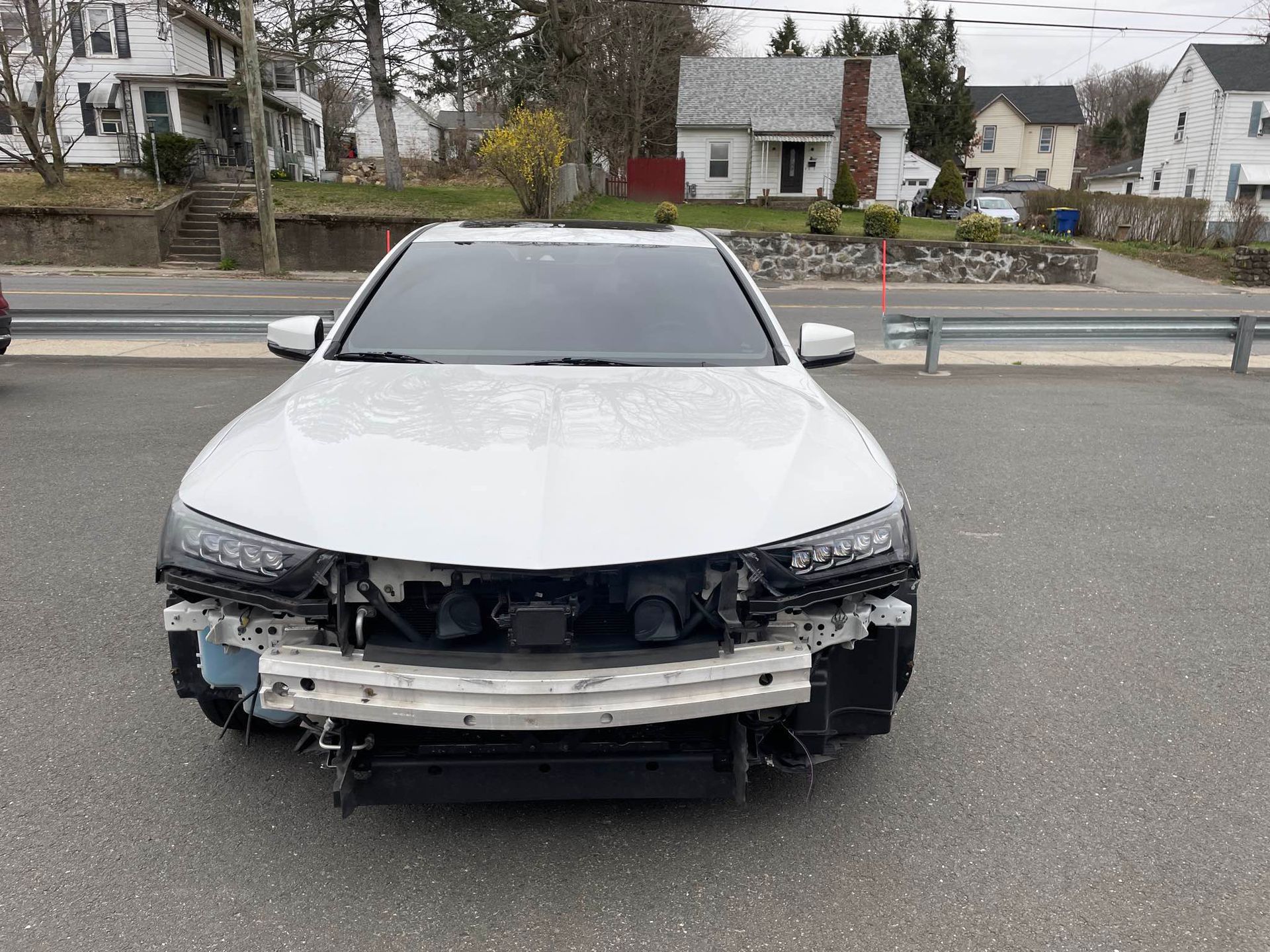 A white car with a broken bumper is parked on the side of the road.