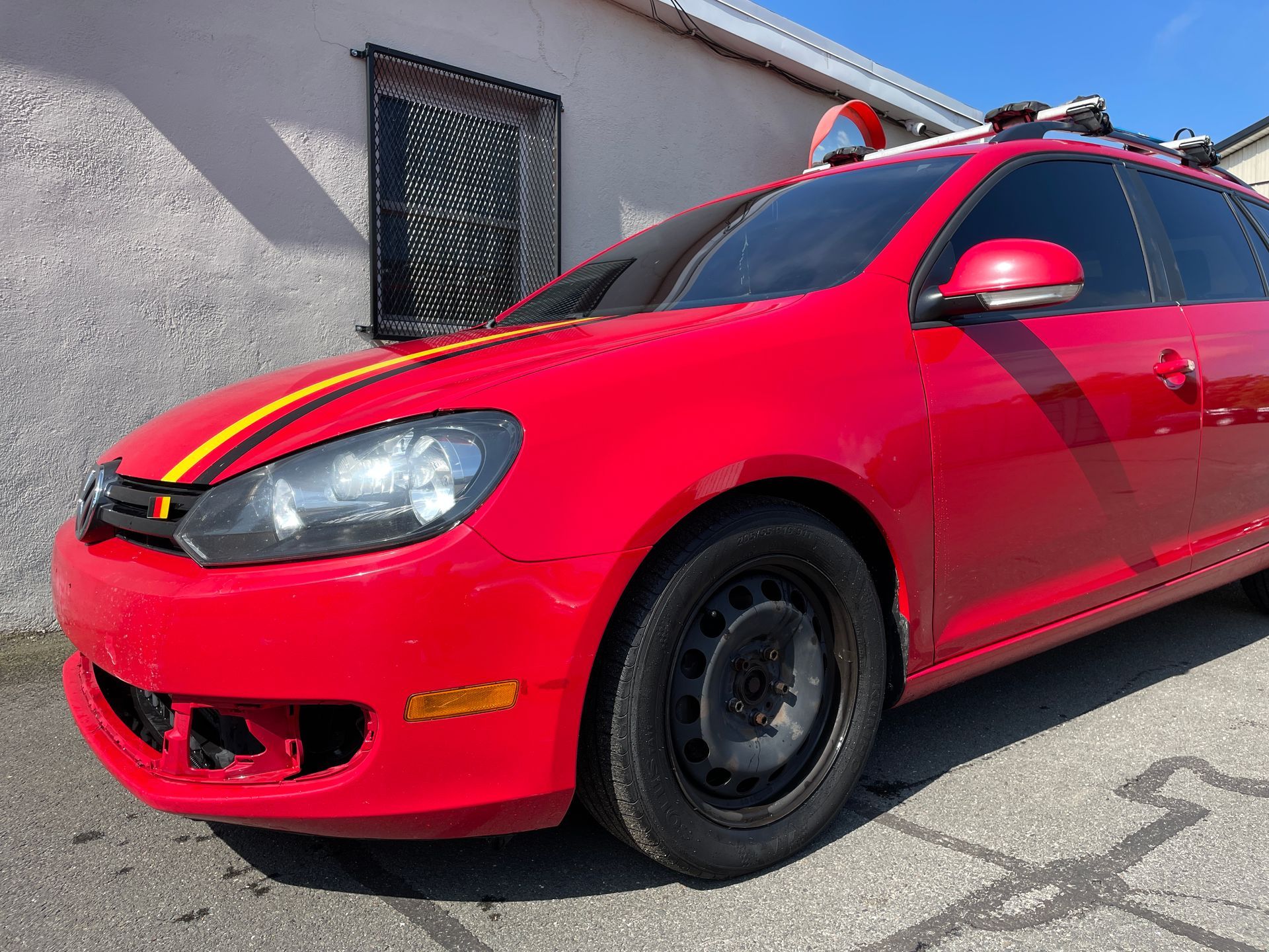 A red car with a yellow stripe on the hood is parked in front of a building.