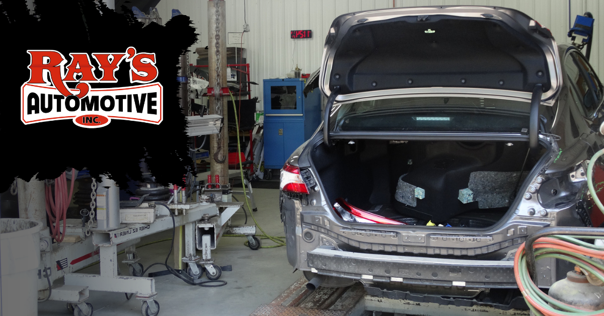 A dark car with its trunk open in a professional automotive repair shop, featuring metal repair equipment in the foreground.
