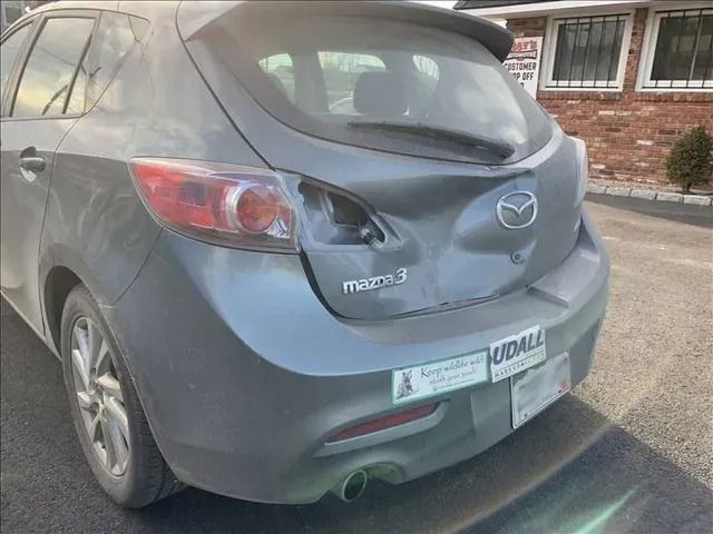A damaged mazda 3 hatchback is parked in front of a brick building.