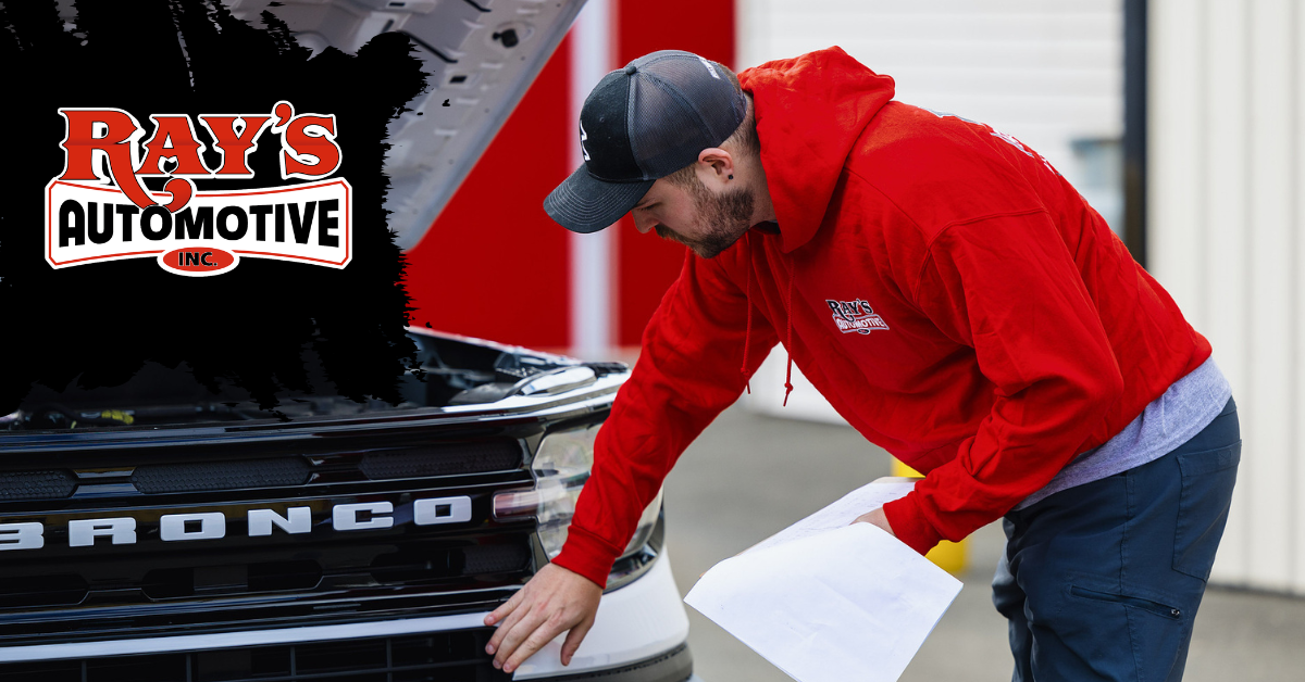 A person in a red hoodie and black cap inspecting the front grille of a Ford Bronco with the hood open, holding papers.