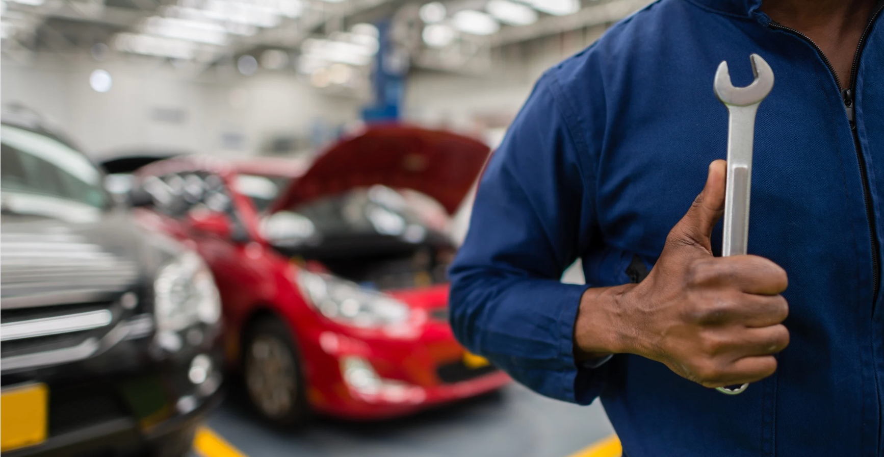 A mechanic is holding a wrench and giving a thumbs up in a garage.