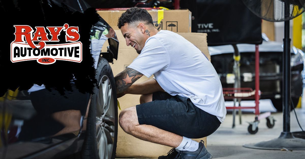 A mechanic in a white t-shirt and shorts crouches to work on a car tire inside a garage with the Ray's Automotive logo.