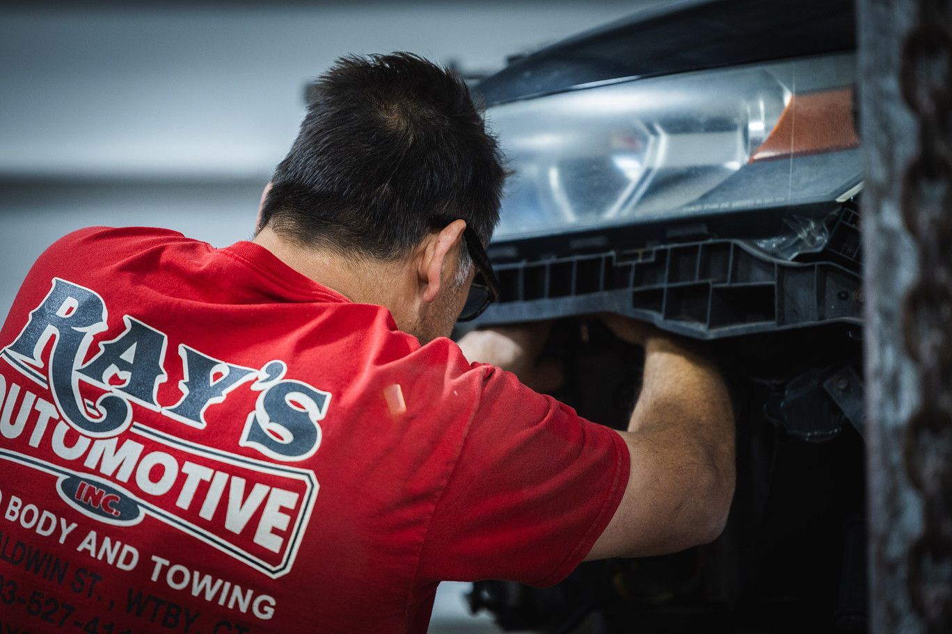 A mechanic wearing a red shirt with Ray's Automotive branding works on the front headlight assembly of a vehicle.