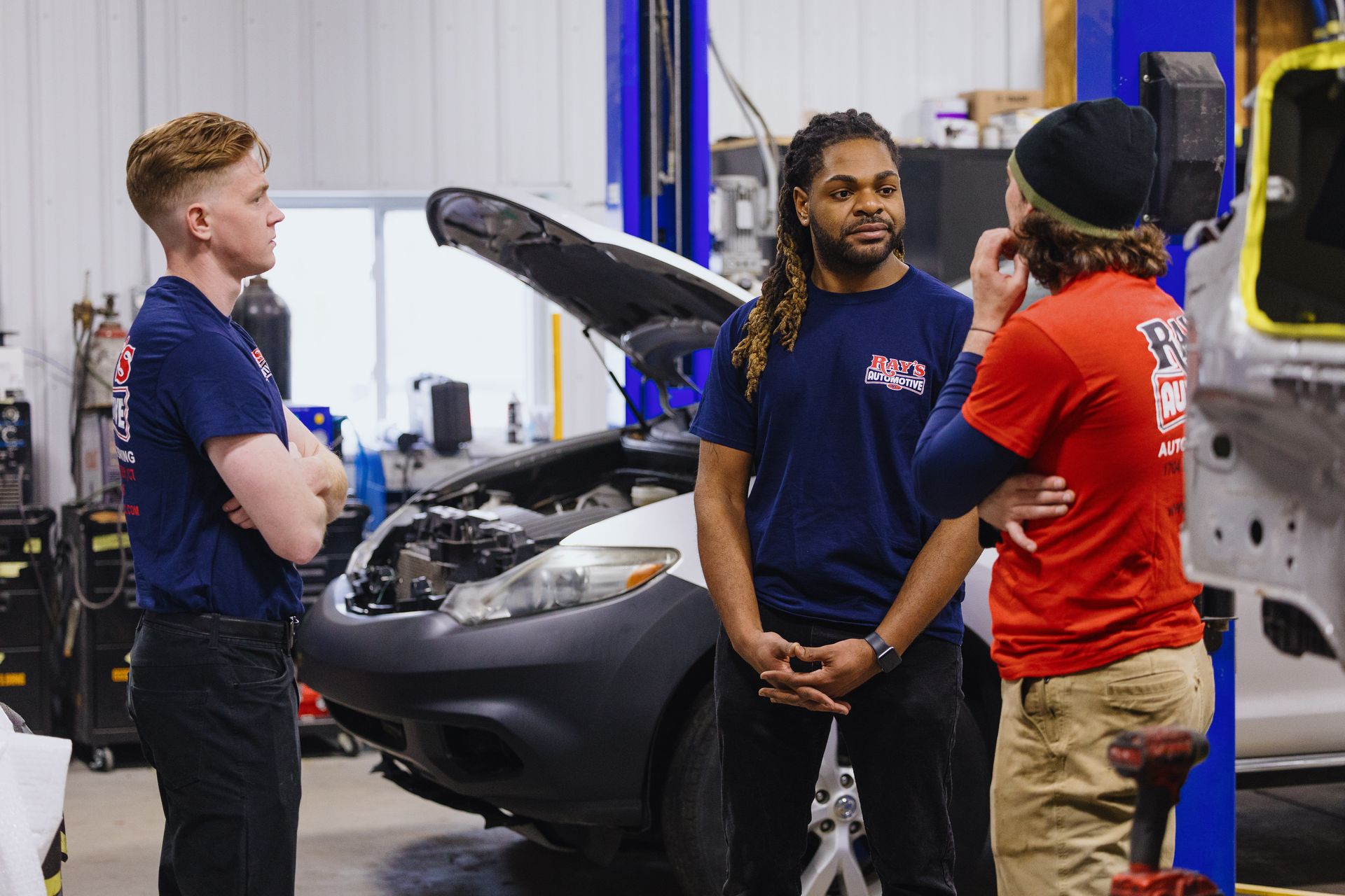 Three people talking beside a car in a mechanic's garage, with the hood open.