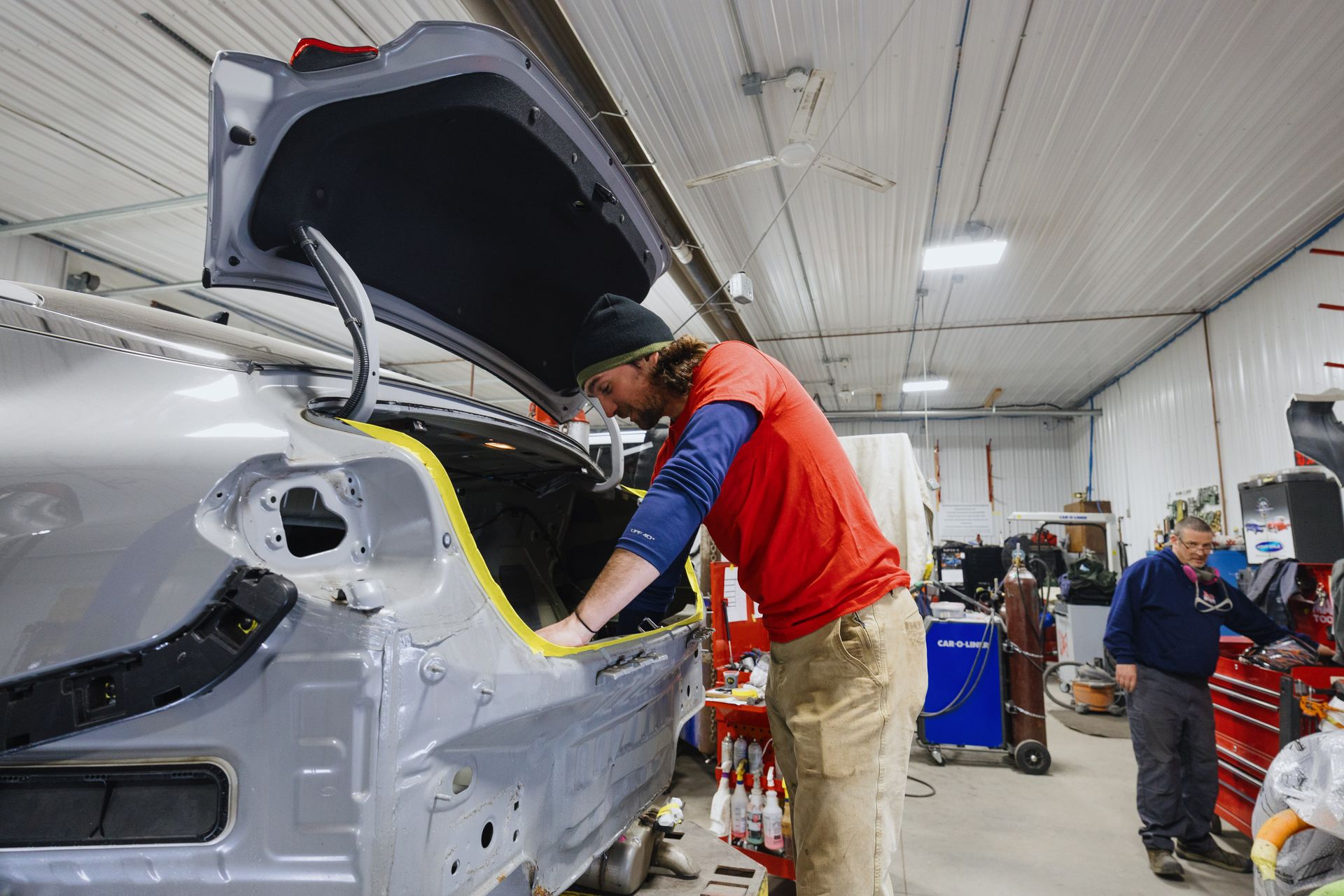 Worker in red vest inspecting a gray car body in an auto repair shop with the trunk open