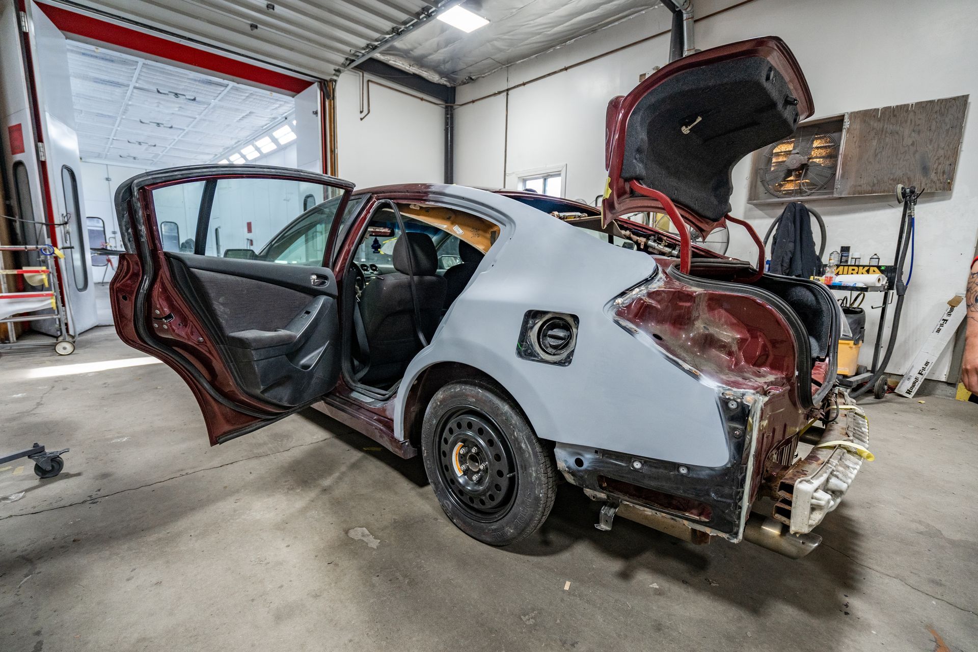 Damaged white sedan in a repair shop with both doors open and the trunk lid raised