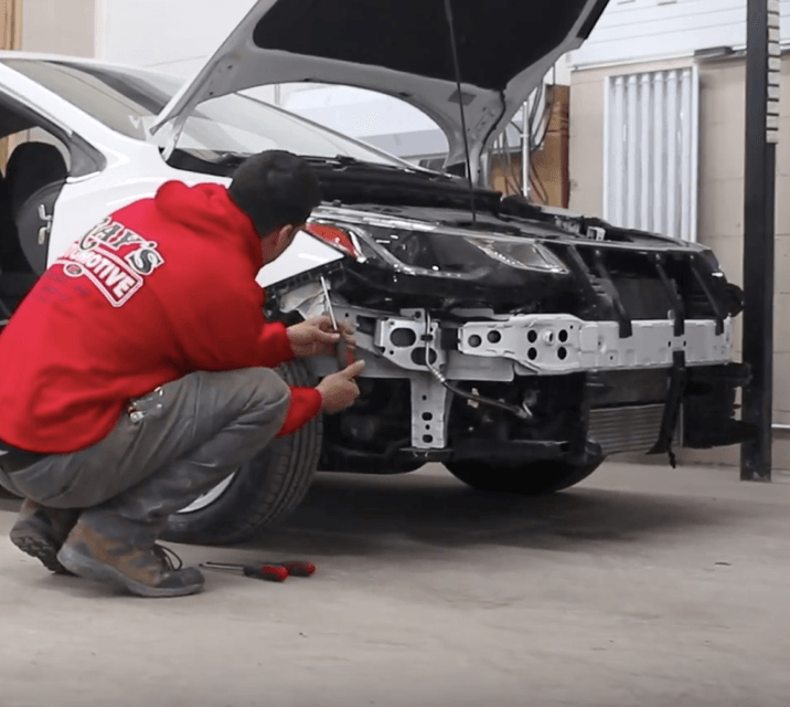 Mechanic in red hoodie repairing a car’s front end with the hood open in a garage