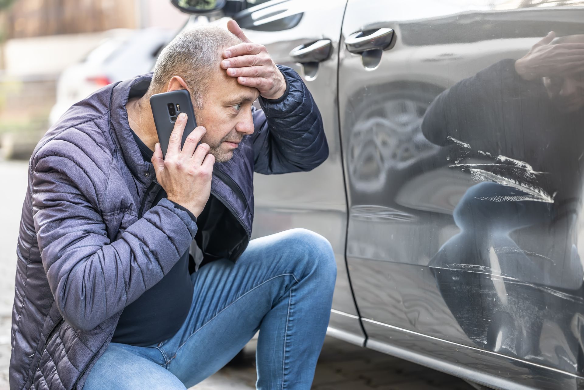 A man is kneeling down next to a damaged car while talking on a cell phone.
