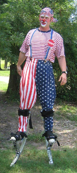 A man dressed as a clown is standing on stilts in the grass.