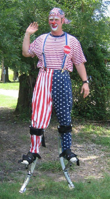 A man dressed as a clown is standing on stilts in a park.