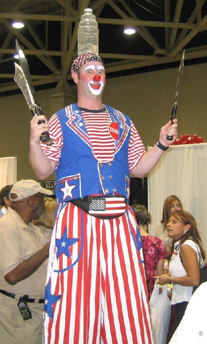 A man dressed as a clown is holding two knives and a bottle on his head