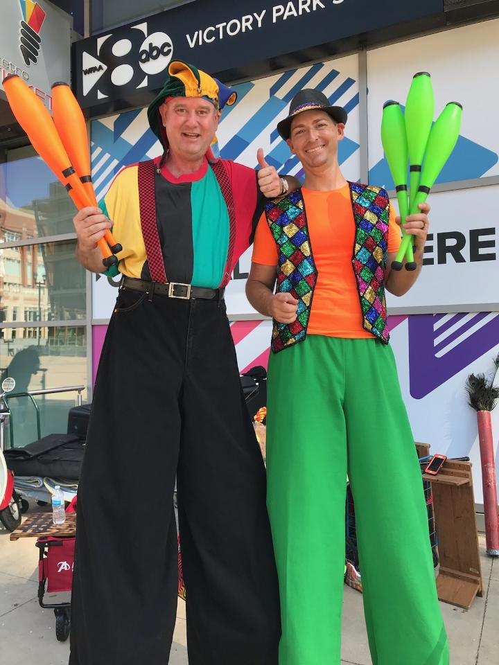 Two men on stilts are holding juggling pins and giving a thumbs up